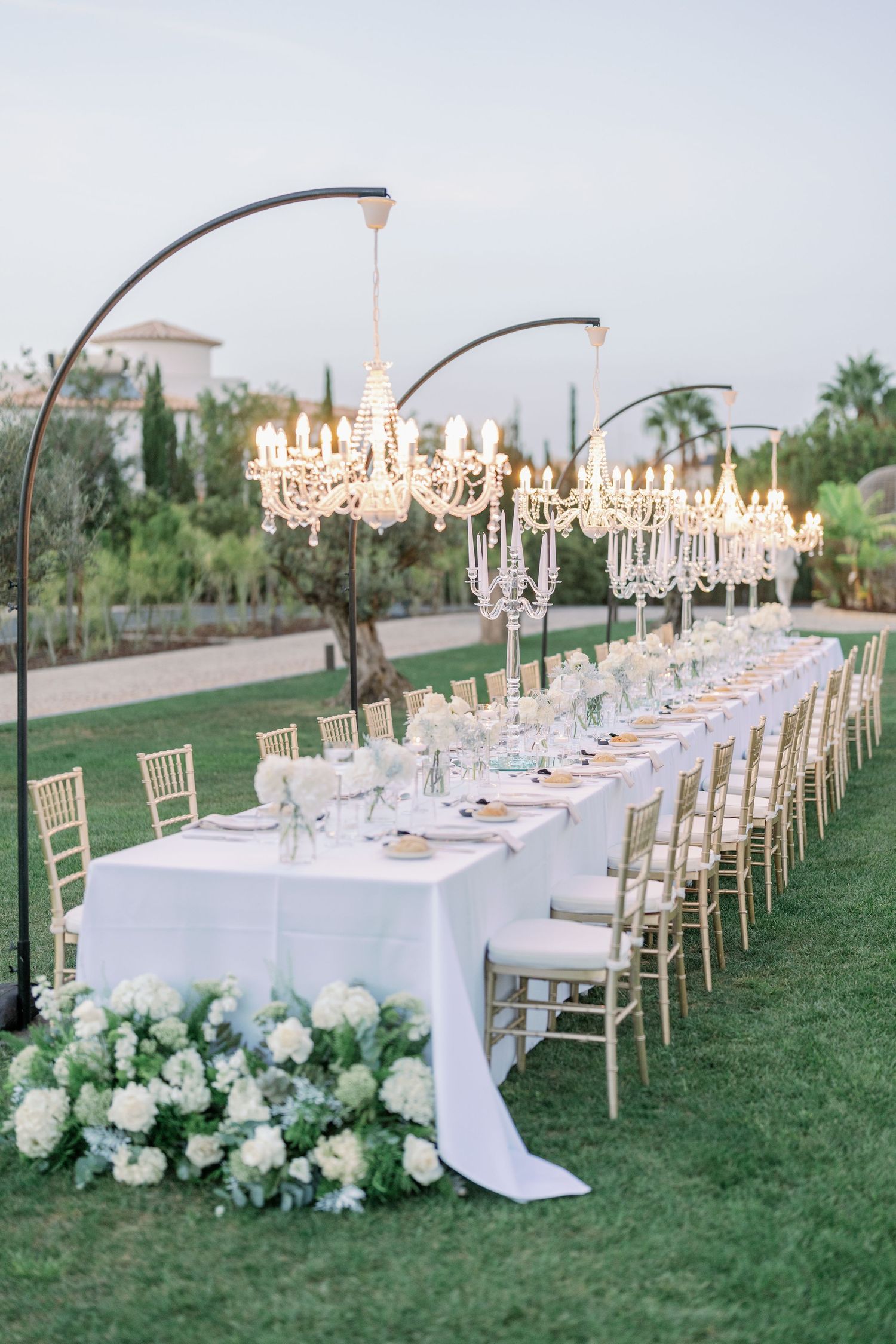 Elegant outdoor wedding reception setup with crystal chandeliers and long white dining tables on grass.