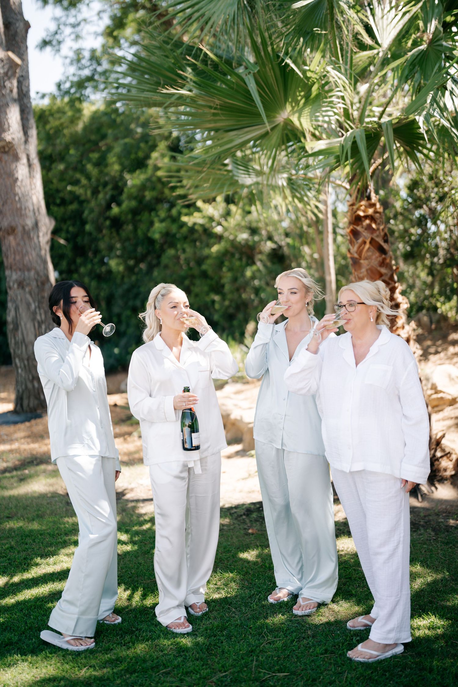 Wedding party in matching white sleepwear poses playfully with champagne under palm trees.