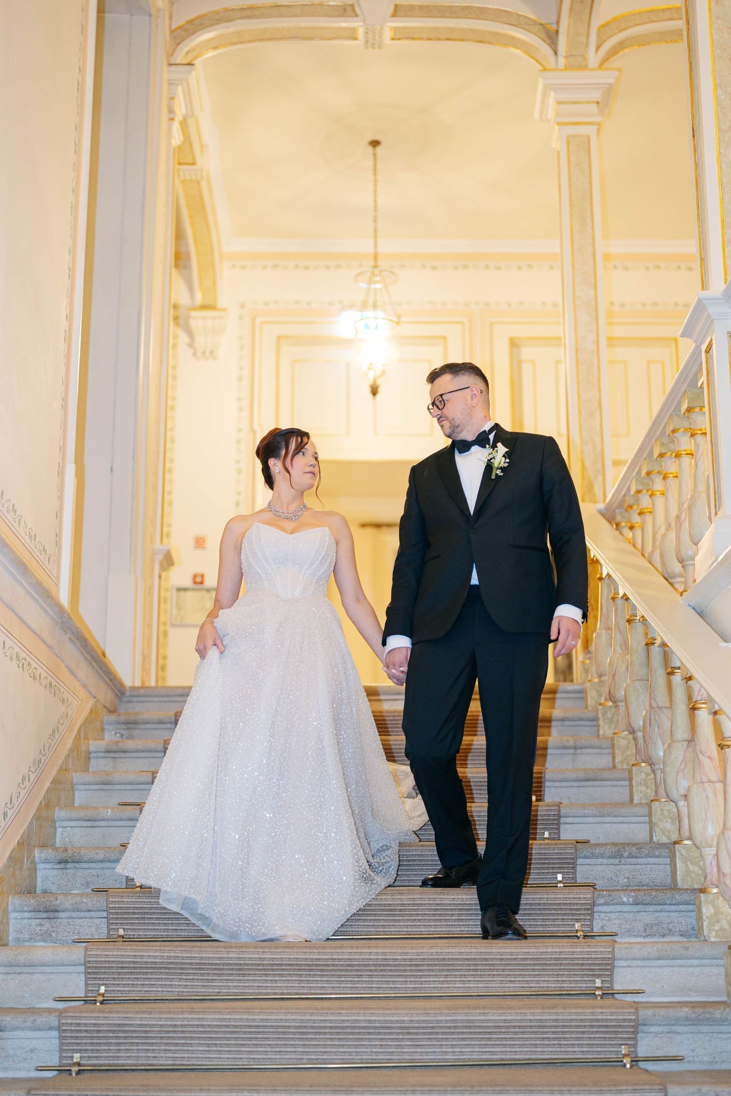Wedding couple walks down elegant white marble staircase in grand venue with multiple romantic poses.