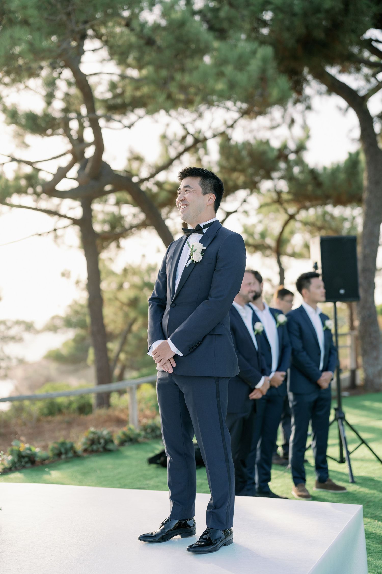 Groom in navy tuxedo standing at outdoor wedding ceremony with groomsmen.