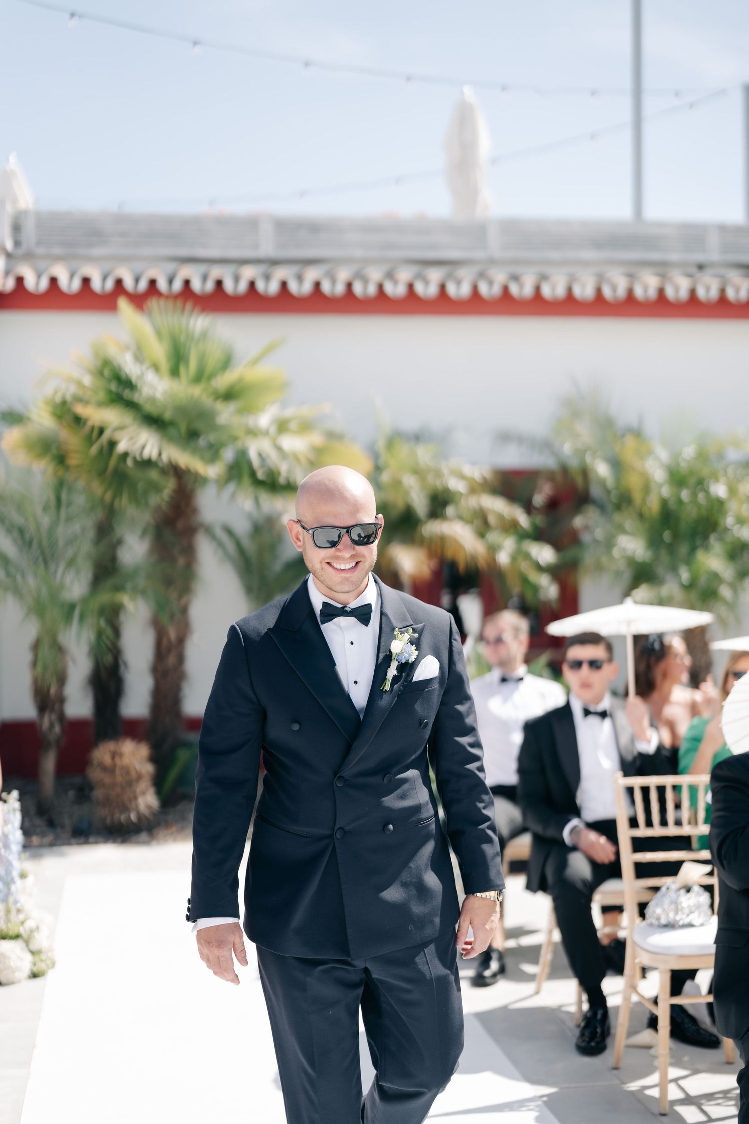 Wedding guests in formal attire walking along a palm-lined venue path.