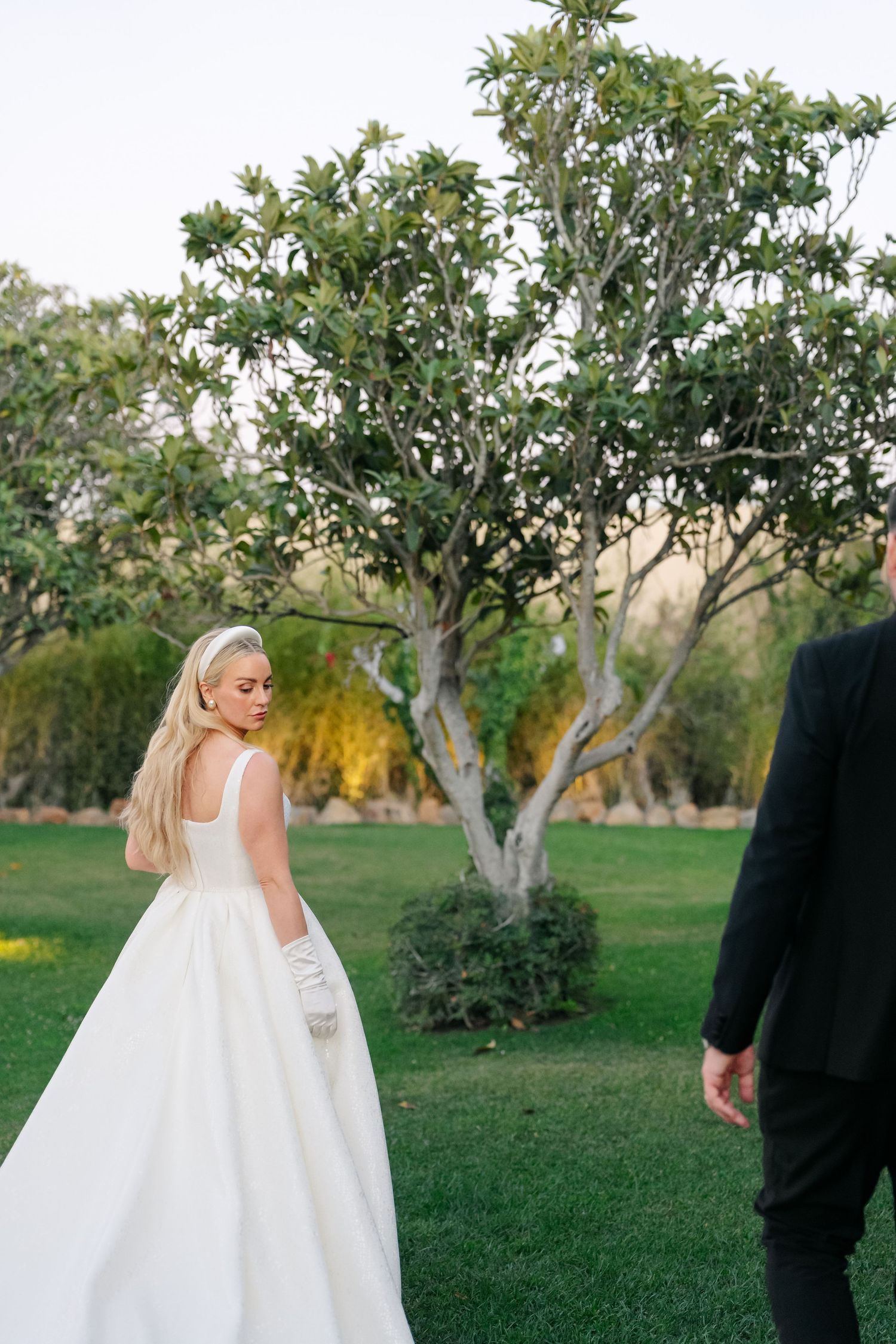 Bride in elegant white gown and groom in black suit explore a garden setting together.