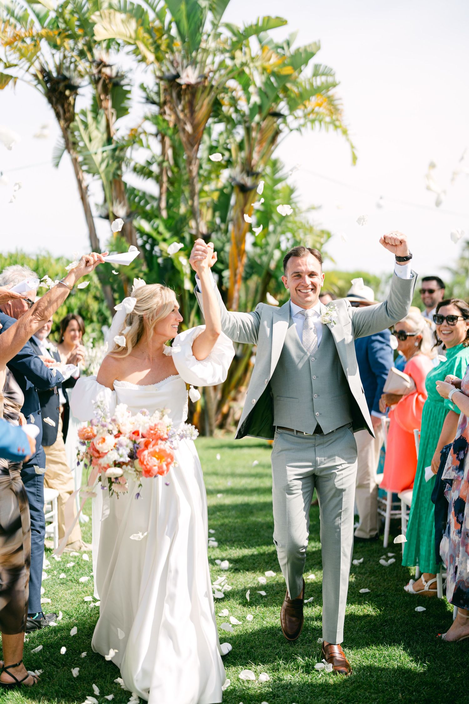Wedding guests and couple celebrate with raised arms during outdoor ceremony under palm trees.