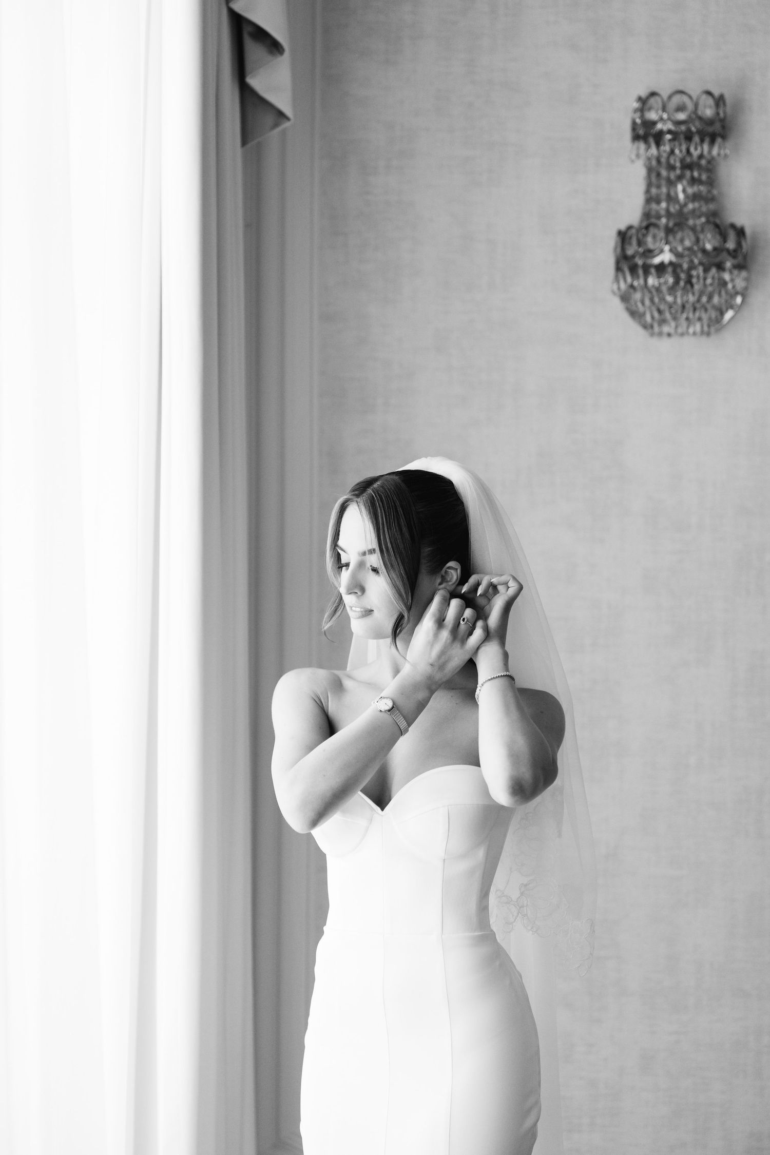 A bride in a white dress poses expressively against a light wall with a chandelier.
