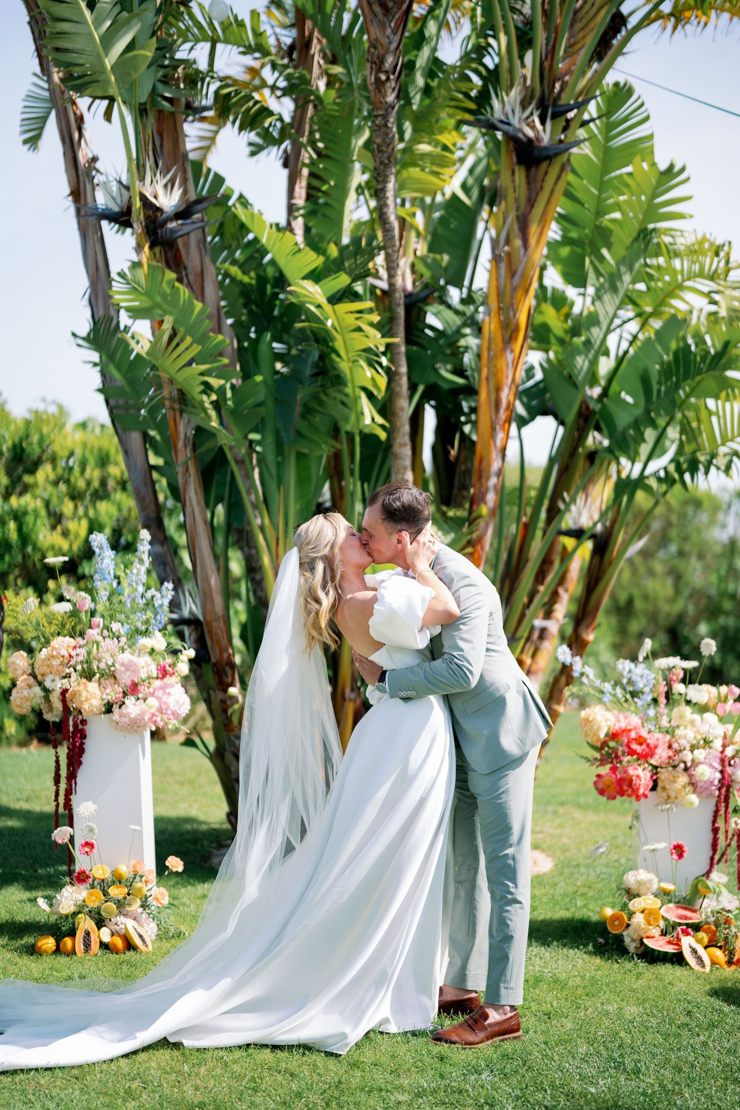 A couple shares a romantic moment in a tropical garden setting with palm trees and colorful floral arrangements.
