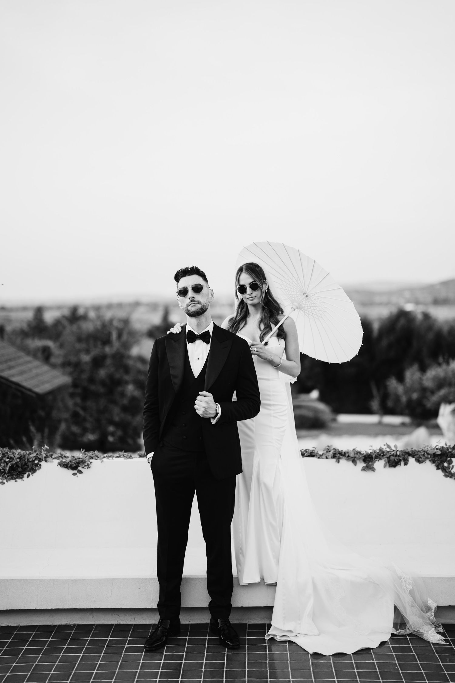Black and white wedding photo series showing couple posing with white parasol on terrace with scenic landscape view.