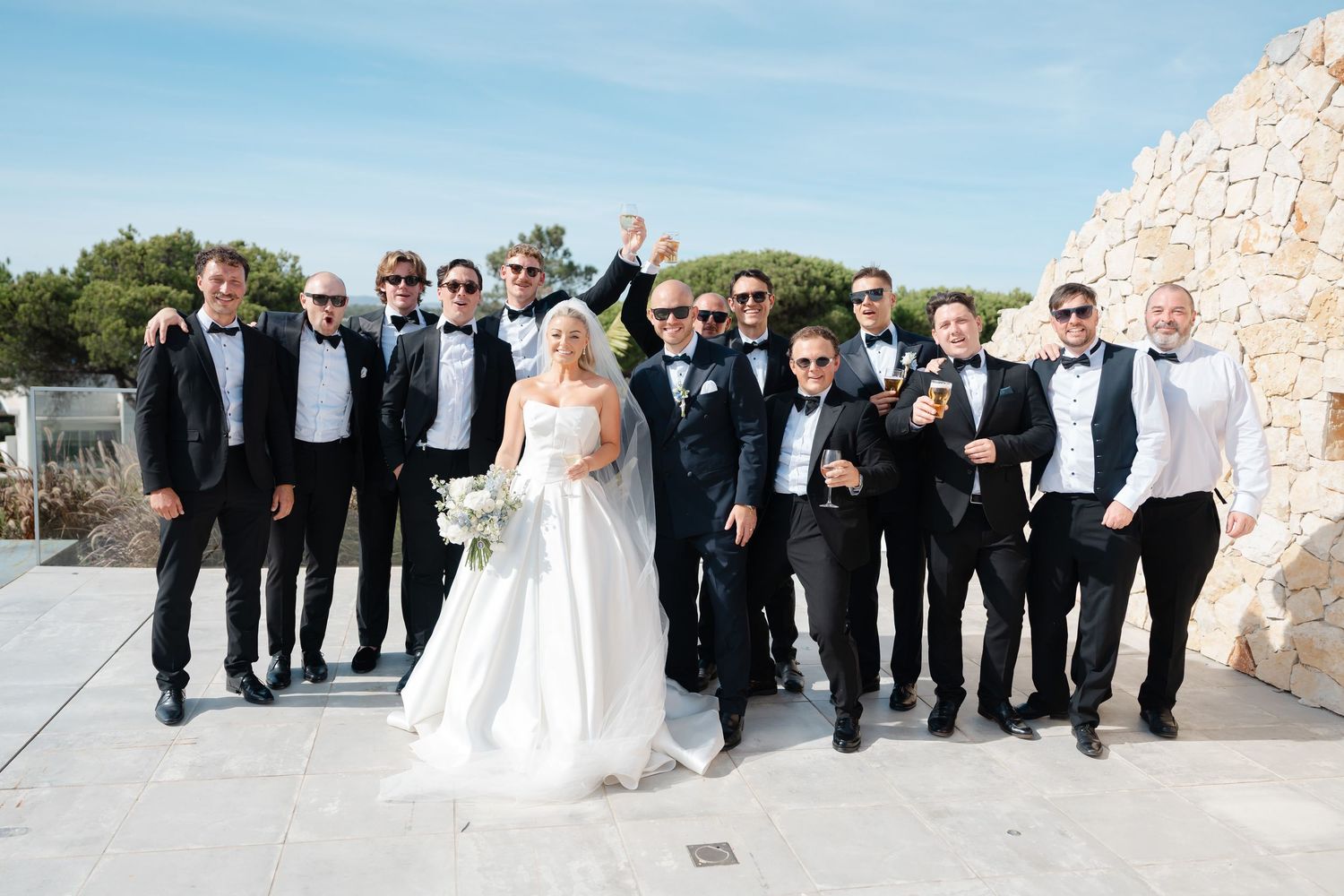 Wedding party in black tuxedos and white dress pose together outdoors.