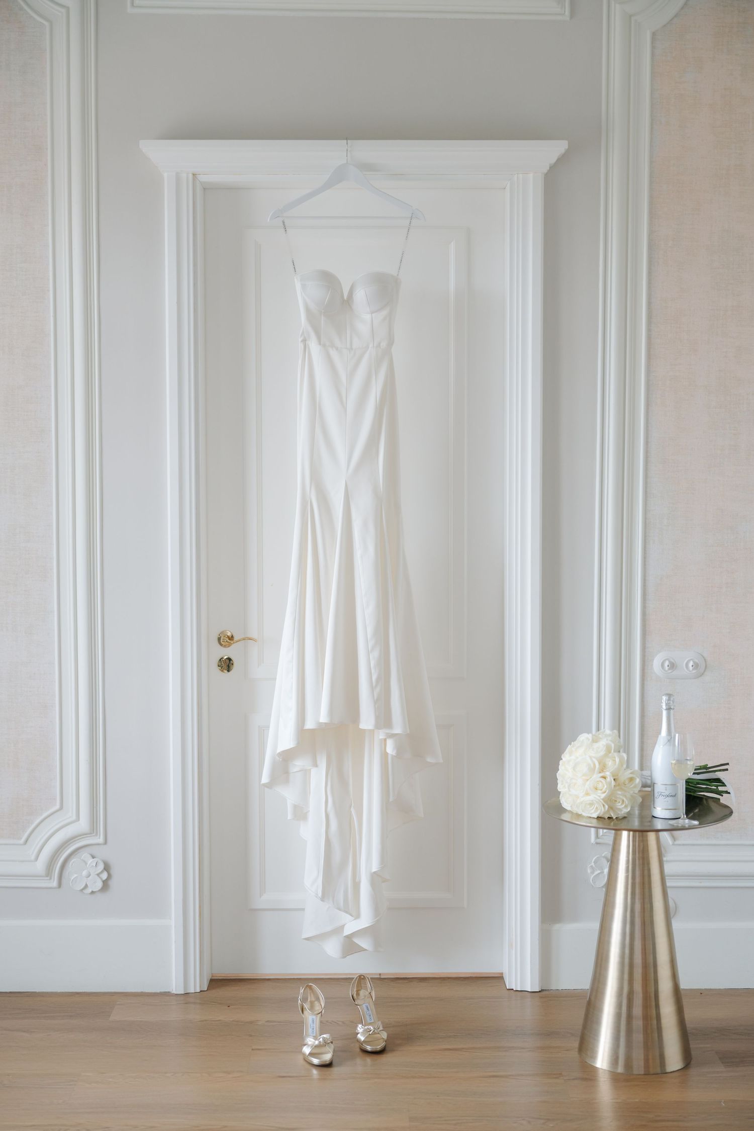 White wedding dress hanging on door with heels beneath and decorative table in elegant room.