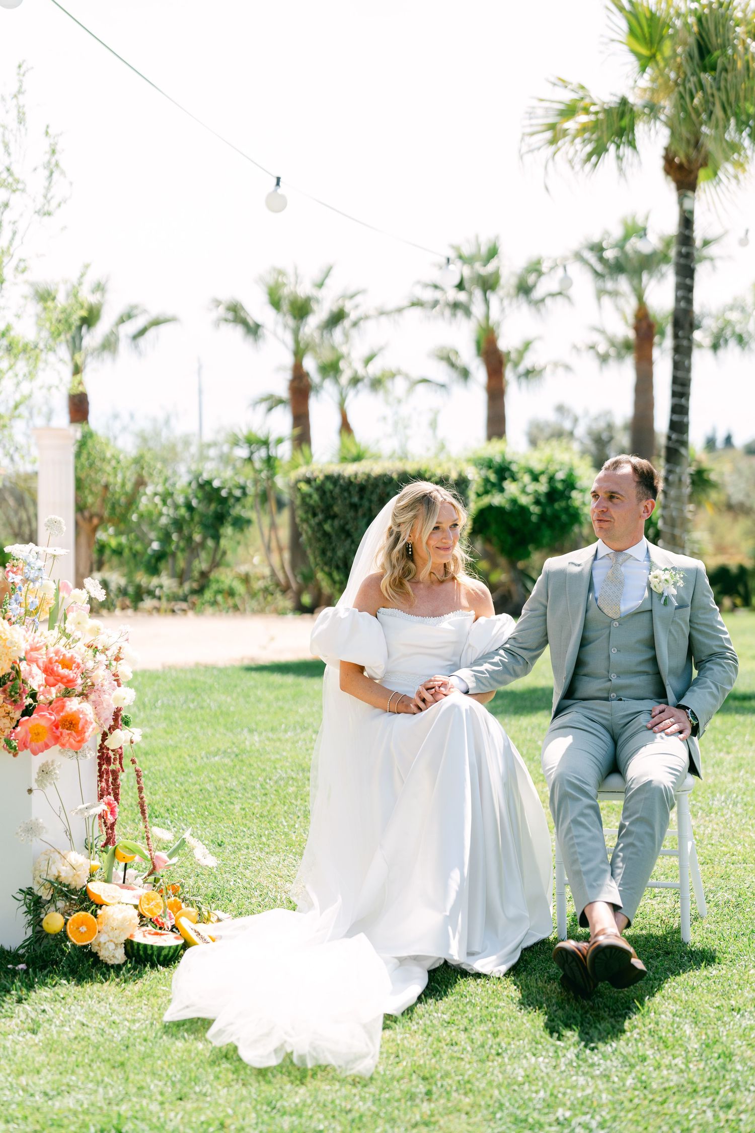 Newlywed couple sits on white chairs in a lush garden setting with palm trees and vibrant floral arrangements.