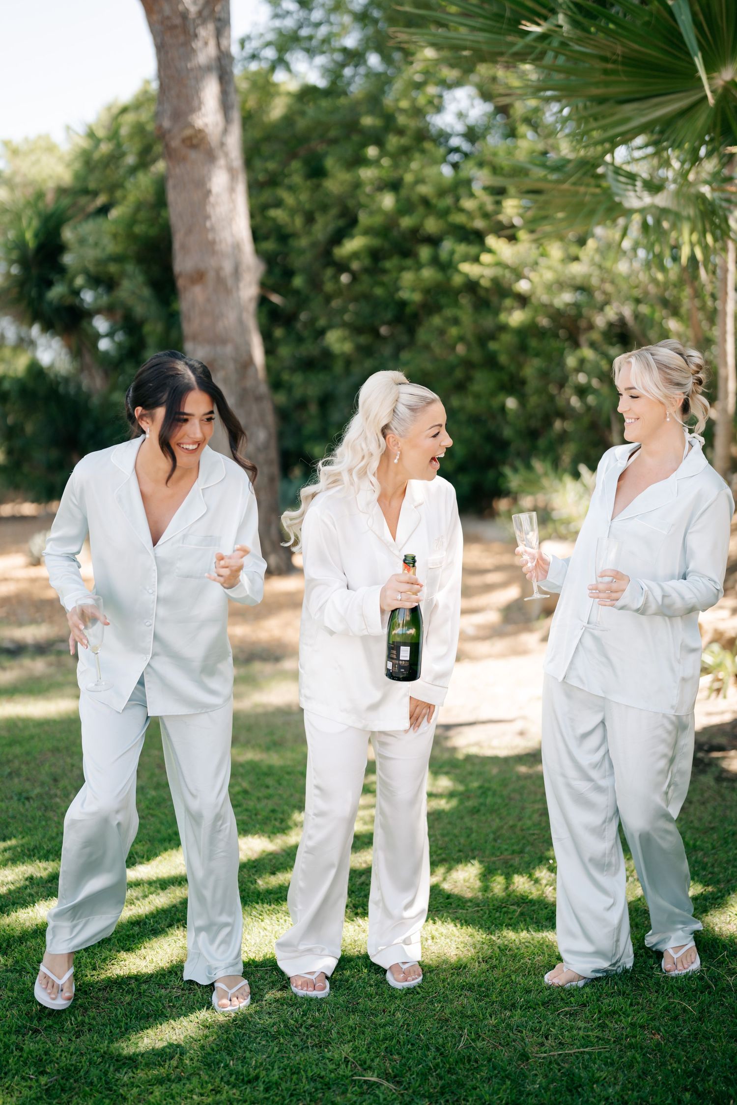 Bridesmaids in matching white silk pajamas celebrate with champagne in a garden setting.