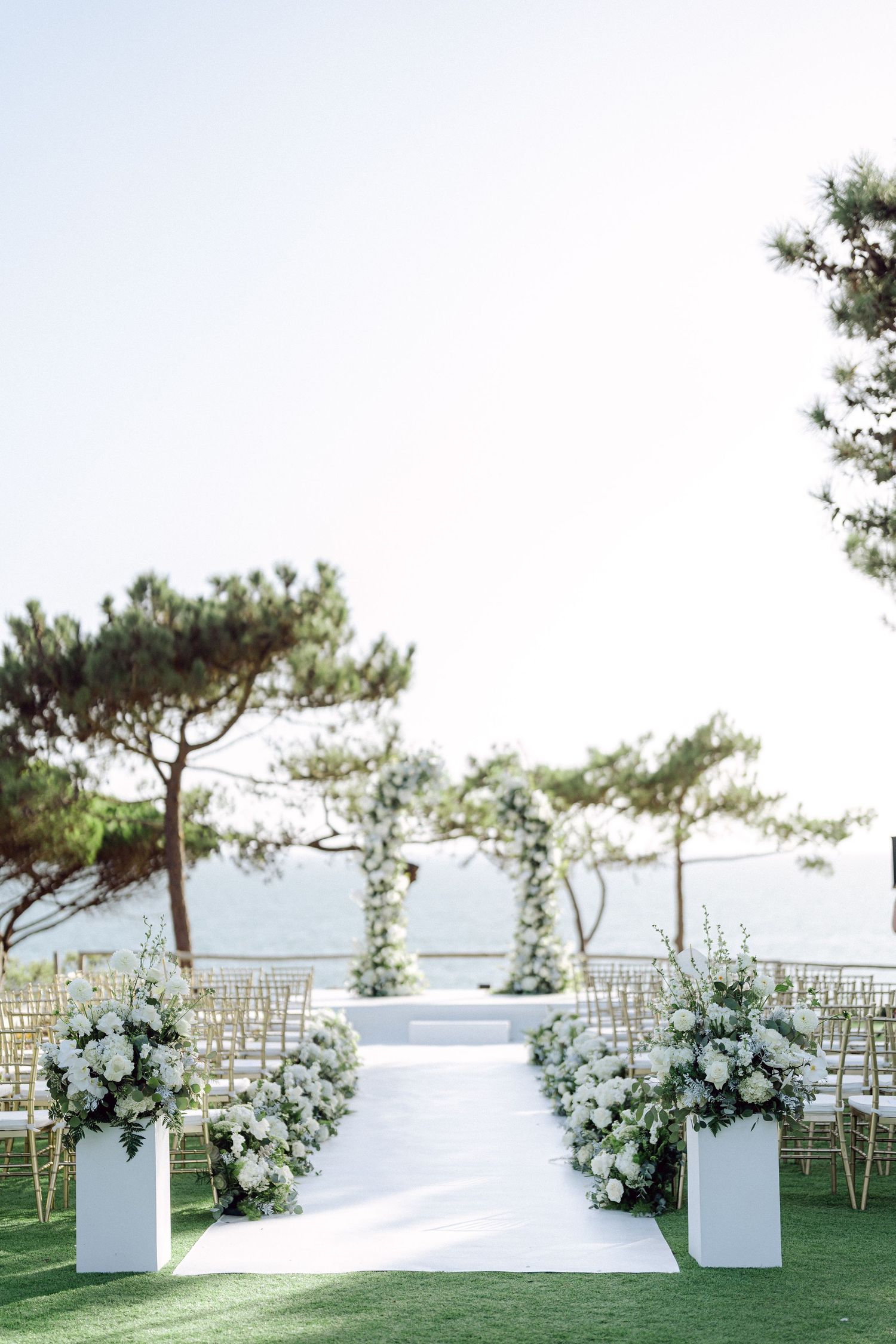 White floral arrangements line an outdoor wedding ceremony aisle with gold chairs and ocean views.