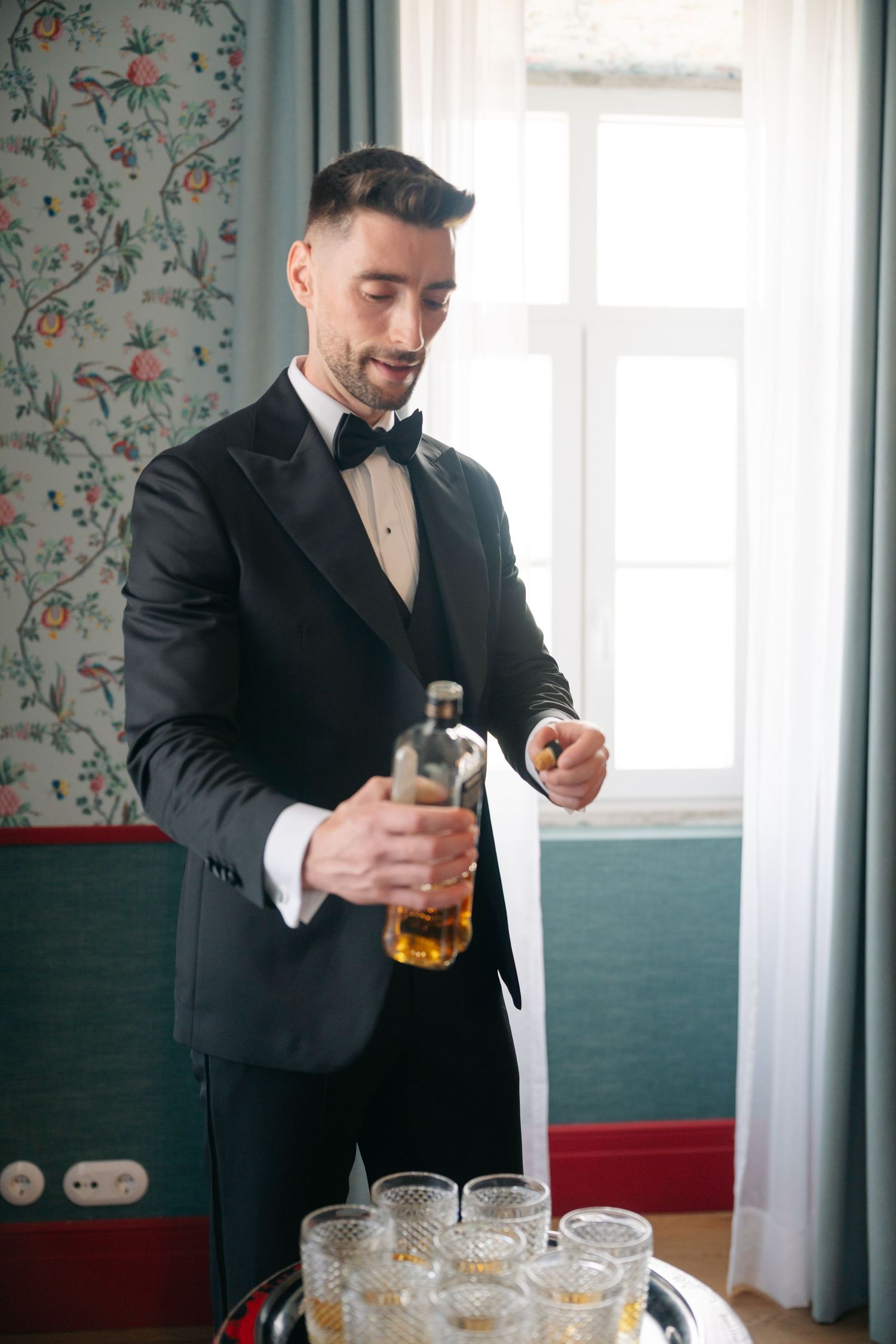 Person in tuxedo pouring drinks from a bottle at an elegant event.