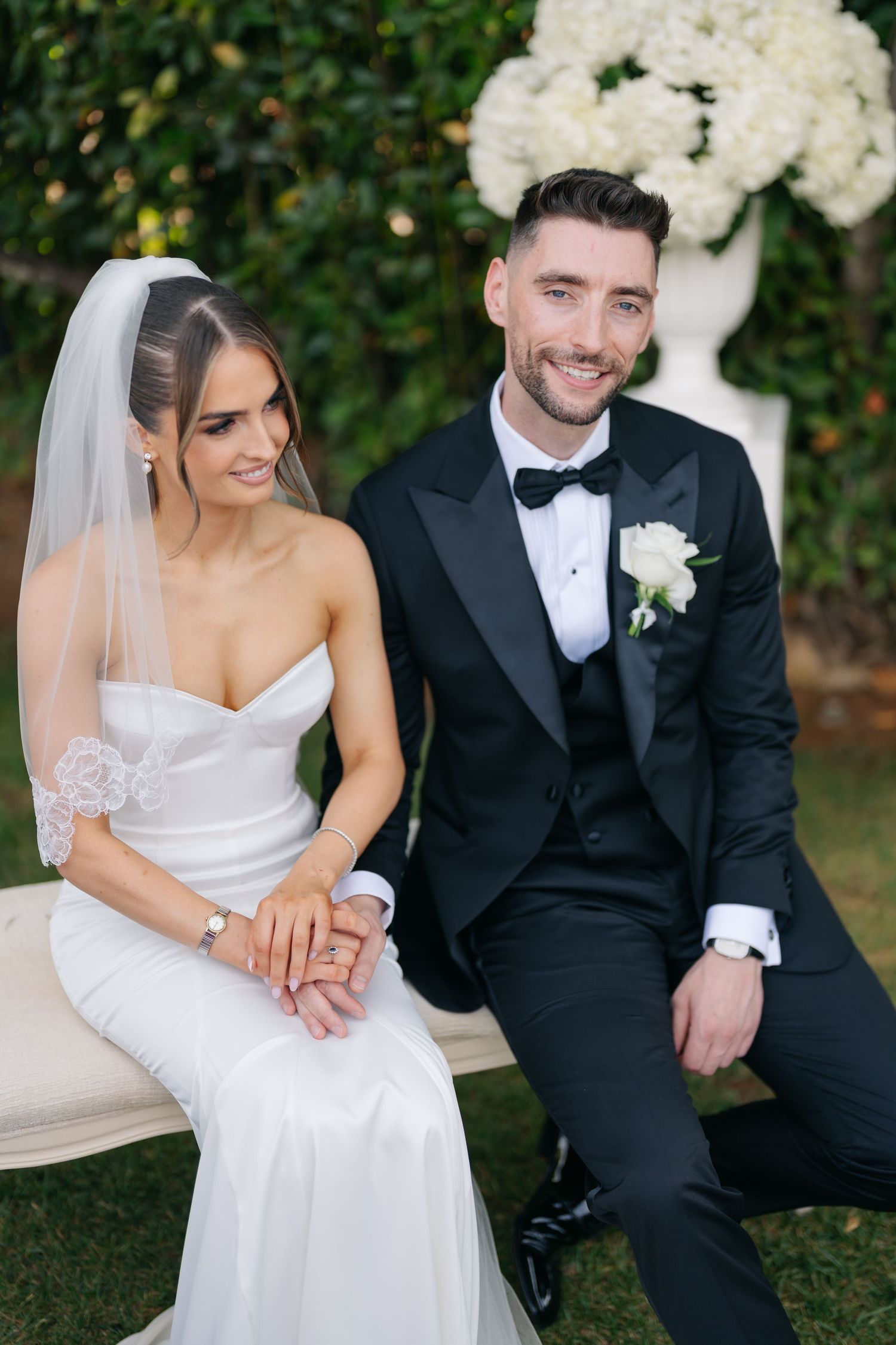 Newlyweds share intimate moments while seated outdoors with white floral arrangements in the background.