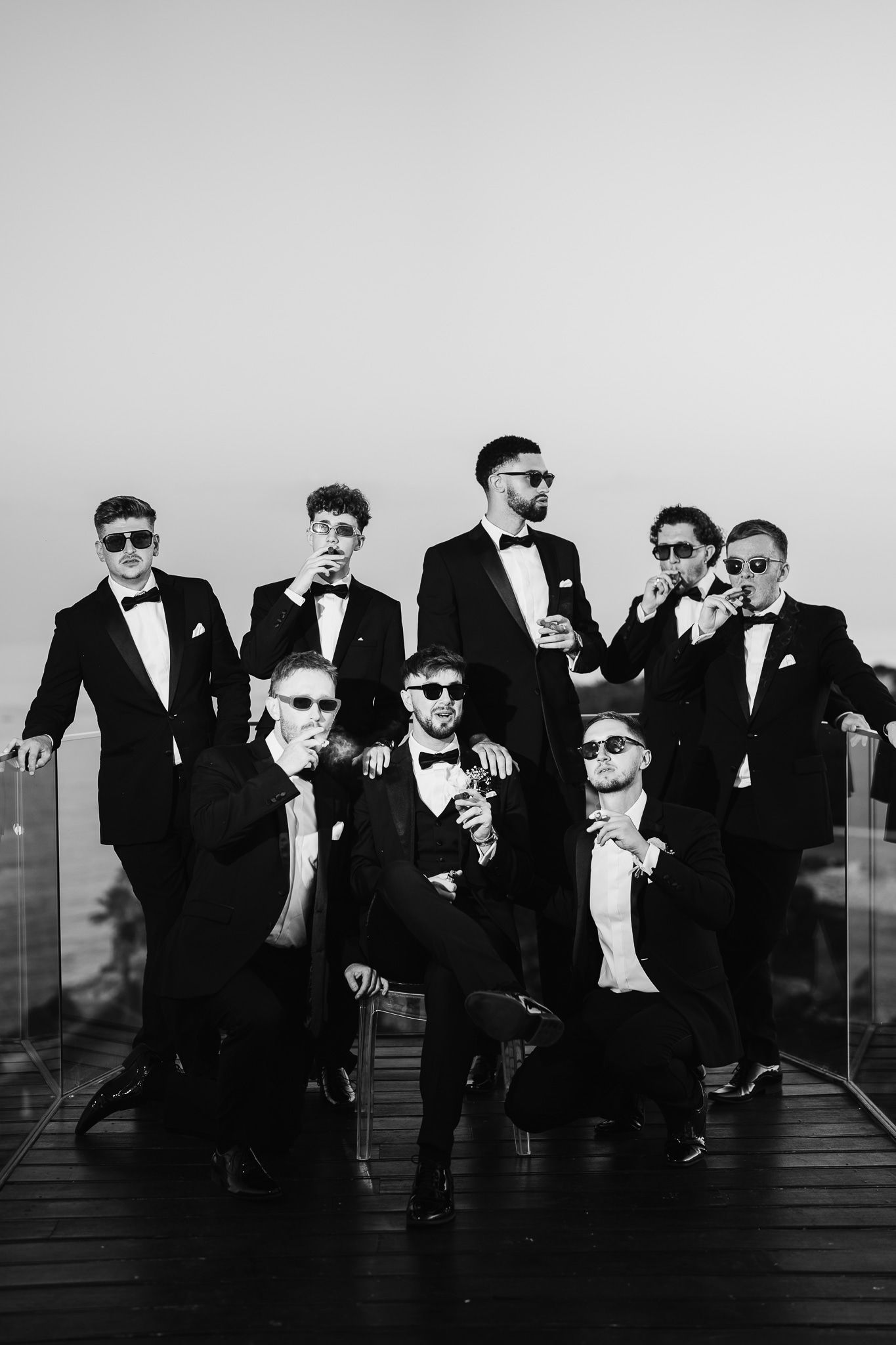 Groomsmen in black tuxedos and sunglasses pose dramatically on outdoor deck.