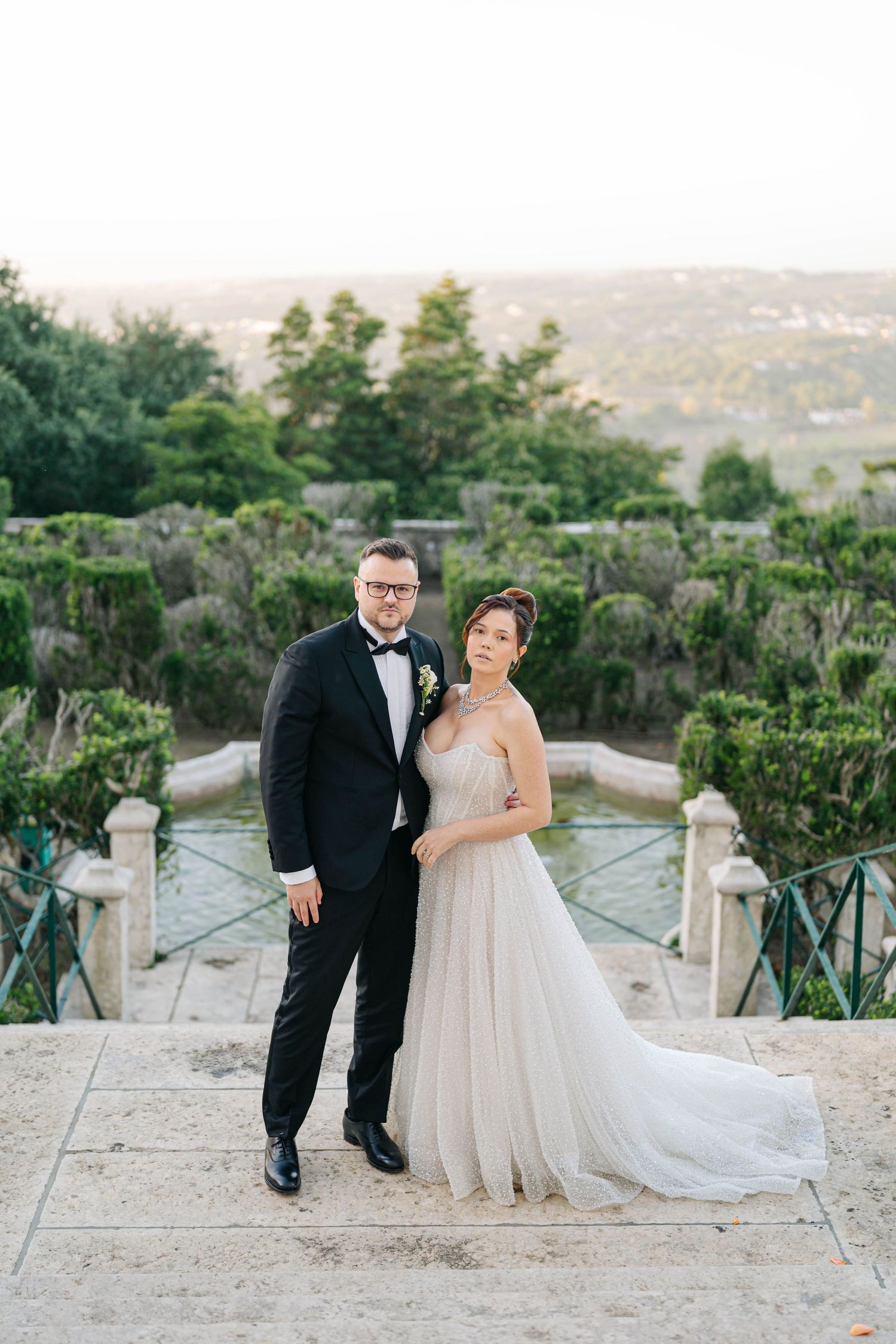 A bride and groom pose together on a stone terrace overlooking a lush garden landscape at dusk.