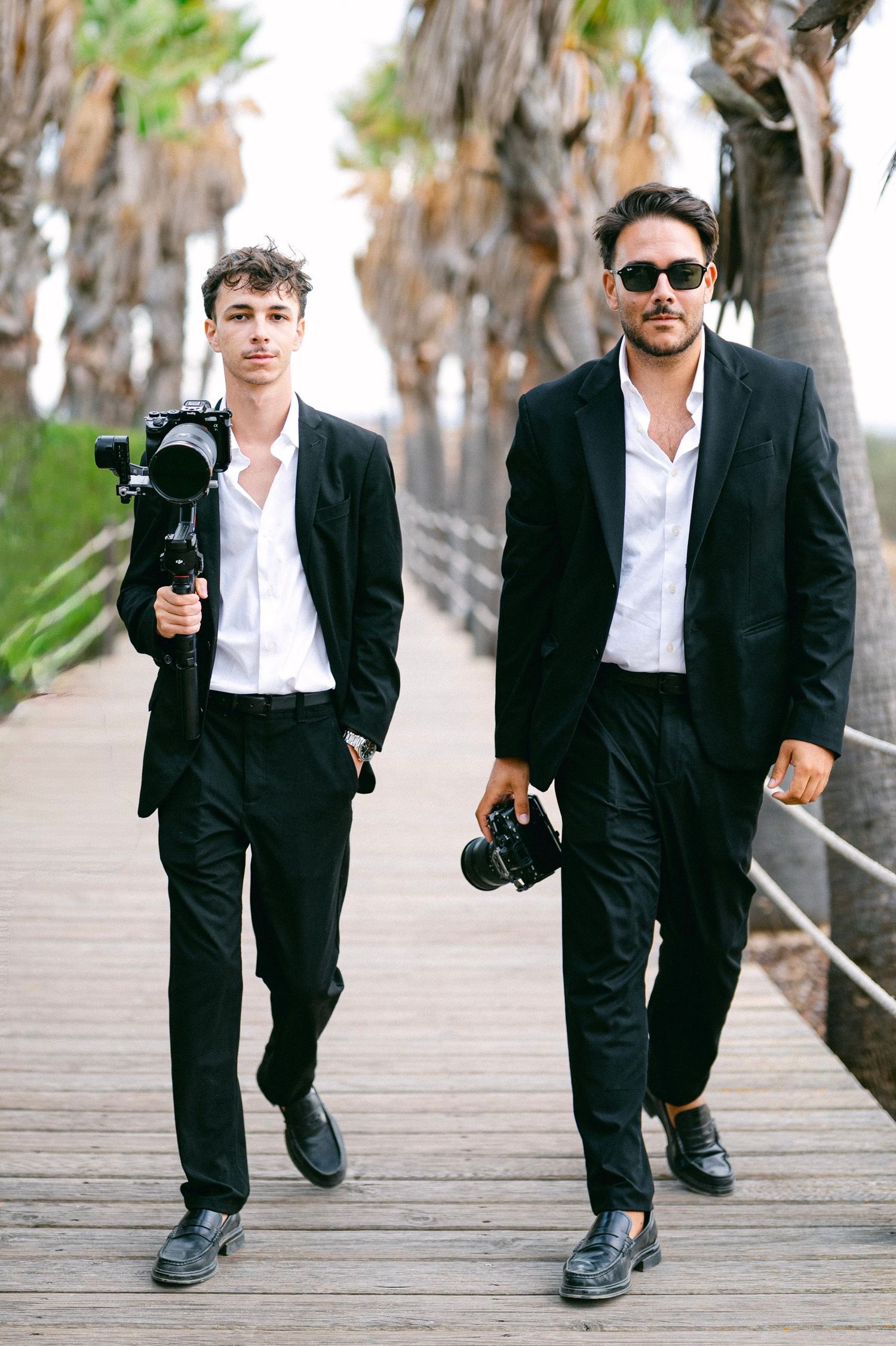 Two professional photographers in black suits walk on a boardwalk carrying cameras, with palm trees in background.