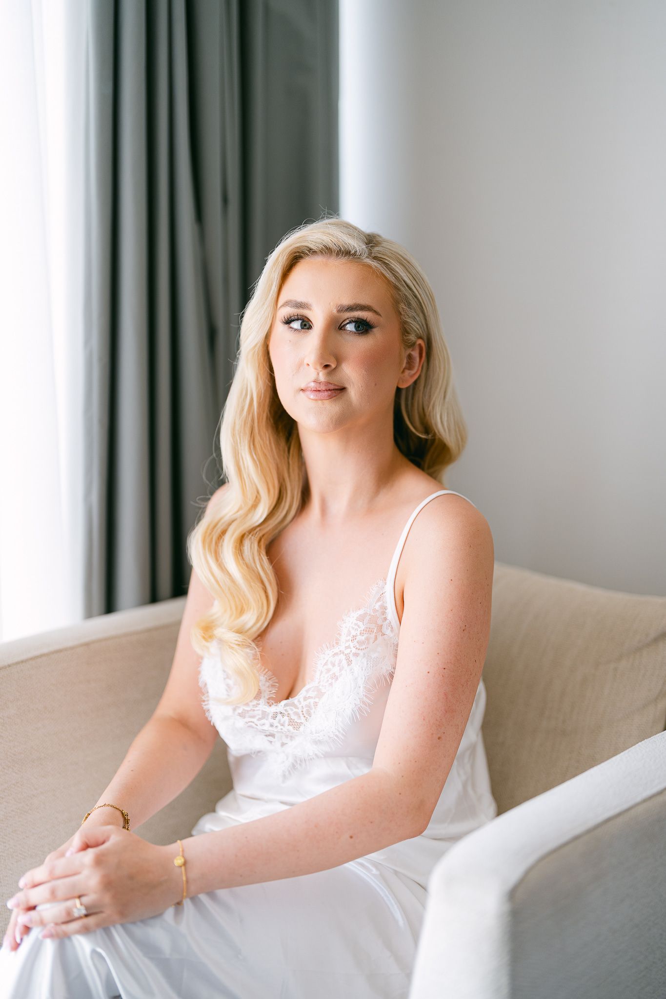 Elegant bridal portrait in white lace dress sitting on beige couch.