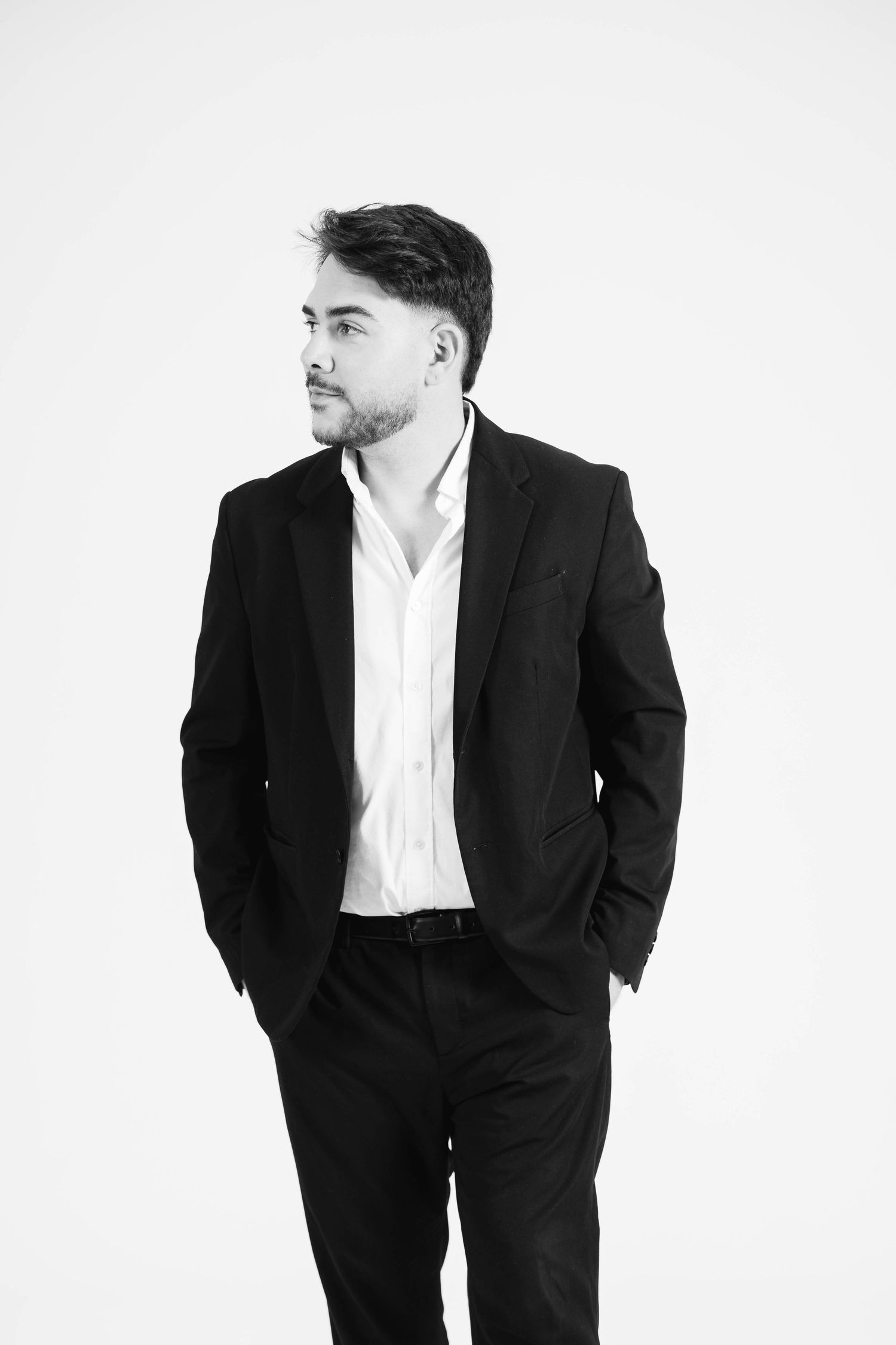 Black and white portrait of a confident man in a dark suit and white shirt, hands in pockets, looking to the side.