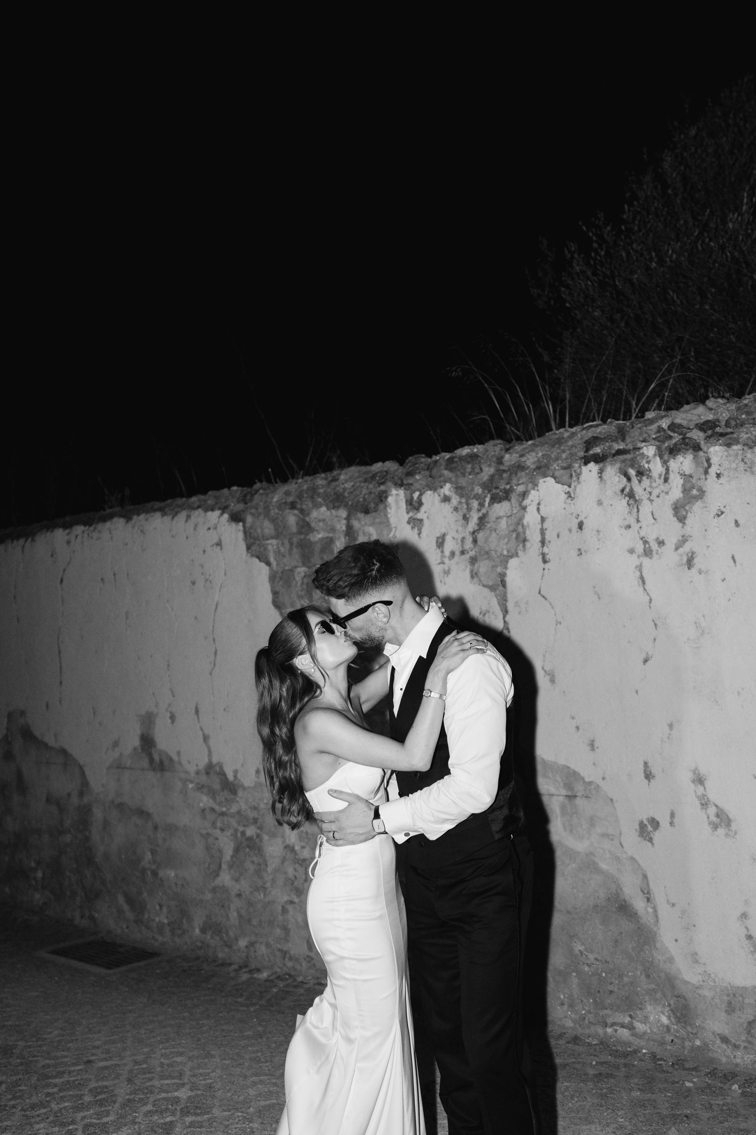A romantic black and white portrait against a textured wall at night.