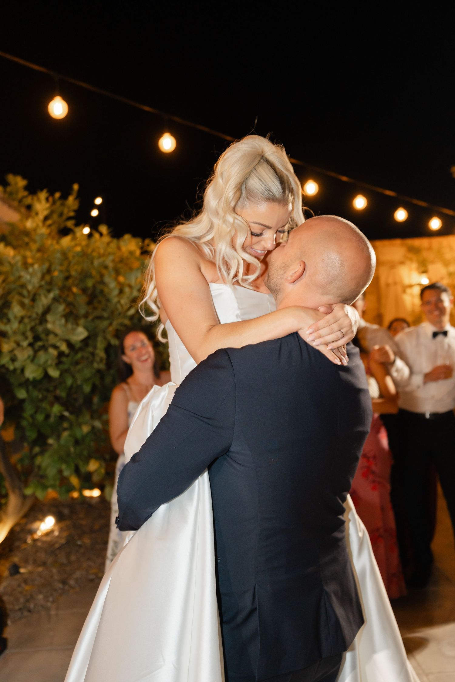 Couple shares an intimate dance moment under warm outdoor string lights at their wedding reception.