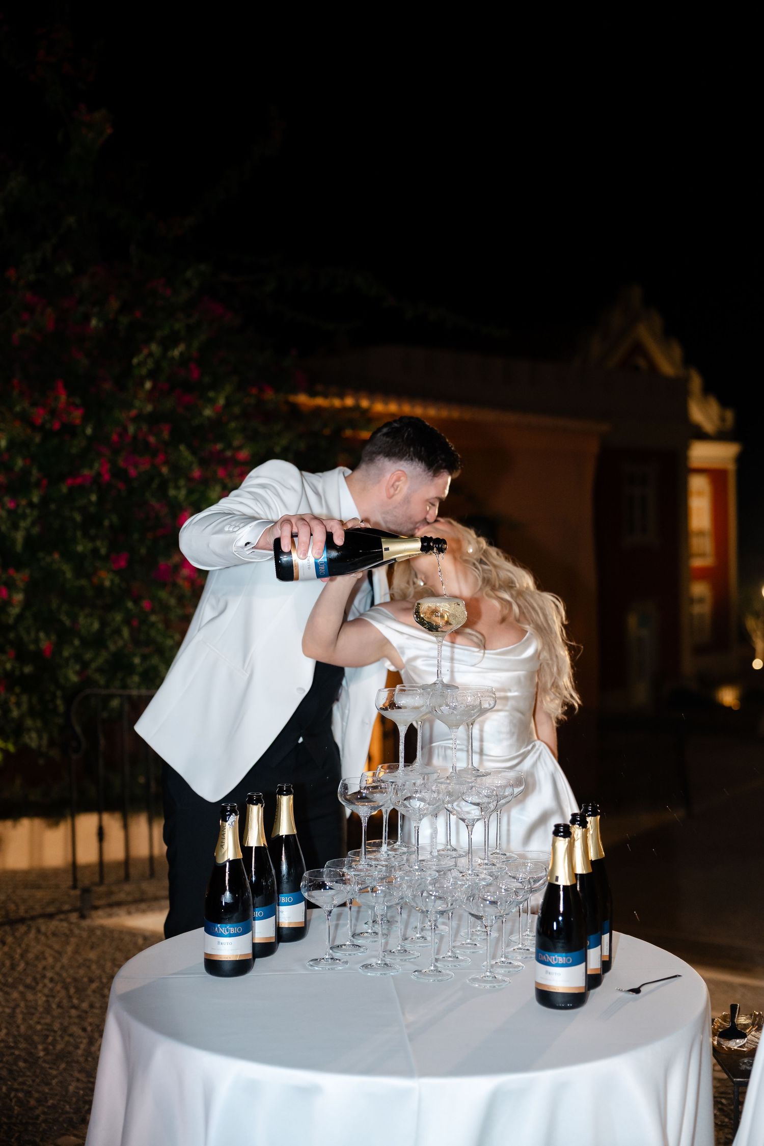 Couple pouring champagne into a tower of glasses at an elegant outdoor wedding reception.