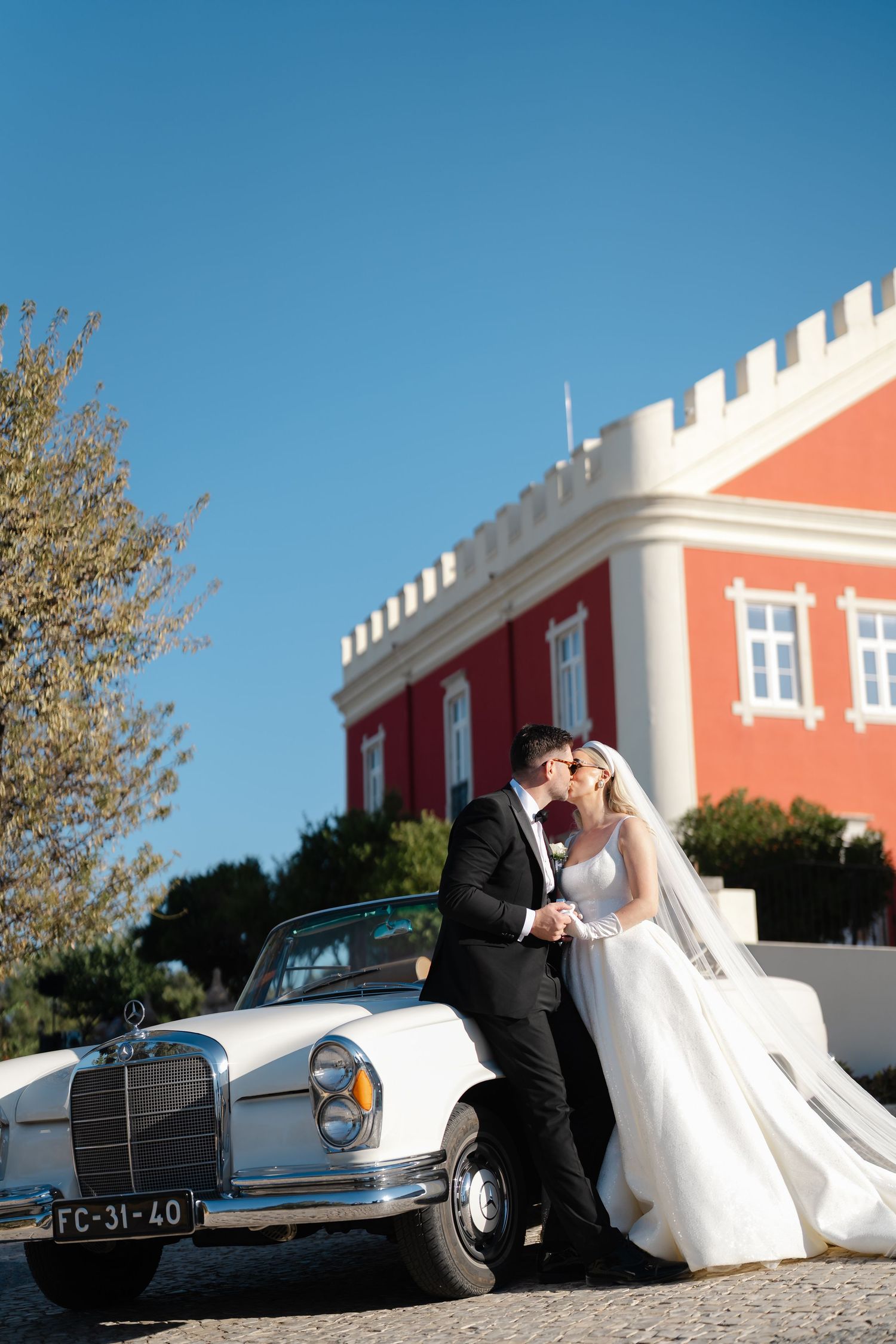 Romantic wedding moment captured against a classic car and coral-colored castle backdrop.