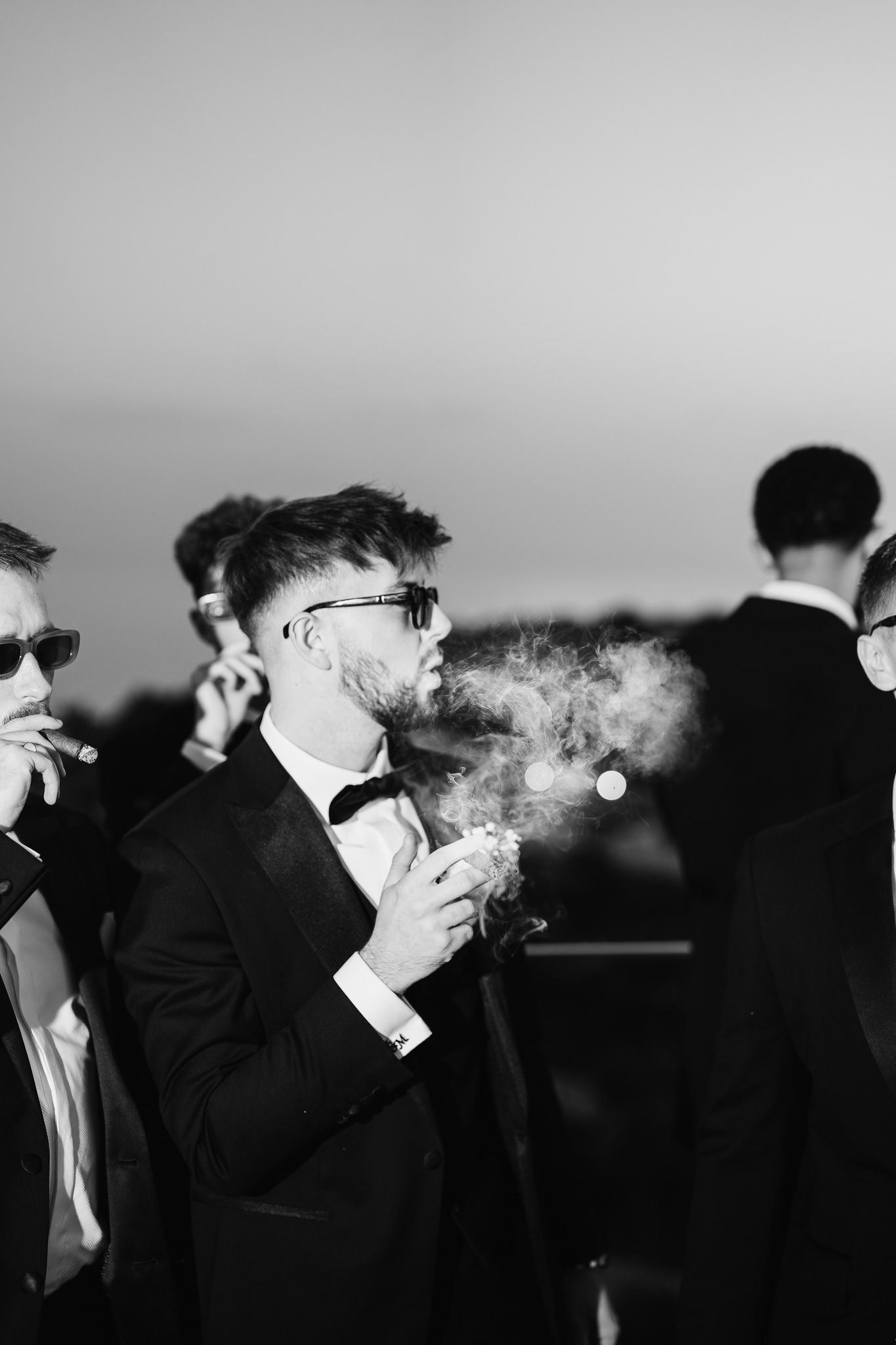 Silhouettes of groomsmen smoking against evening sky in artistic black and white.