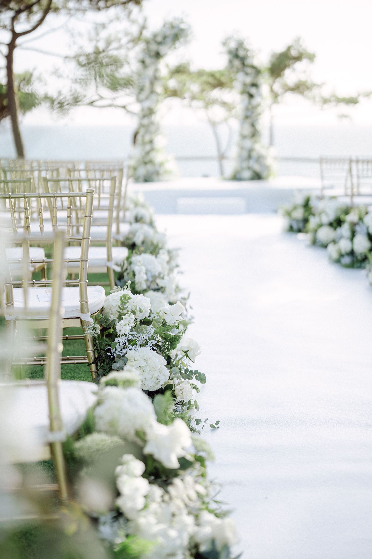 Close-up of white floral aisle decorations with gold chairs at an outdoor wedding venue.
