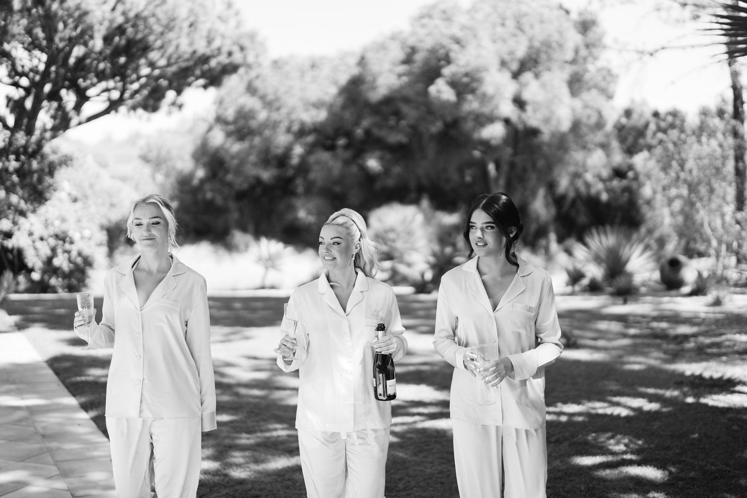 Wedding party members in white silk pajamas celebrate outdoors in natural lighting.