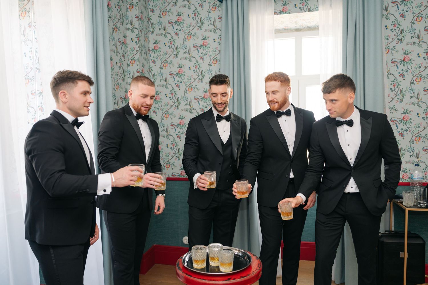 Groomsmen in black tuxedos gathered around drinks at wedding preparation.