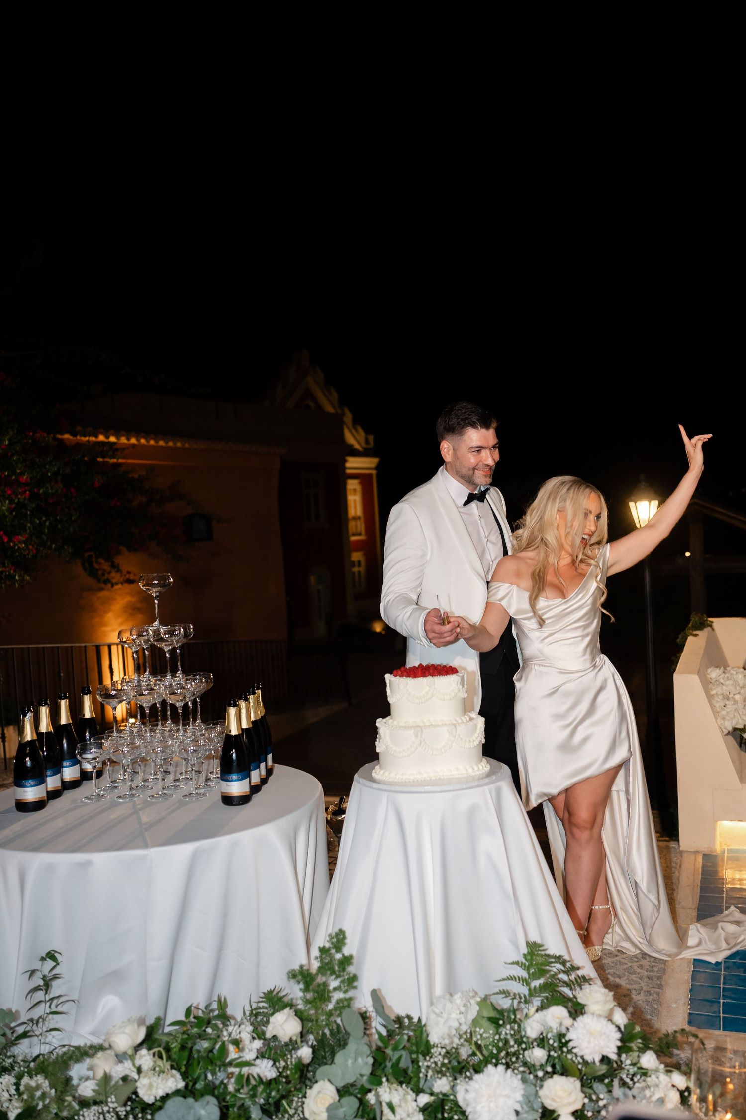 A couple celebrating near tables with champagne and a wedding cake decorated with flowers.
