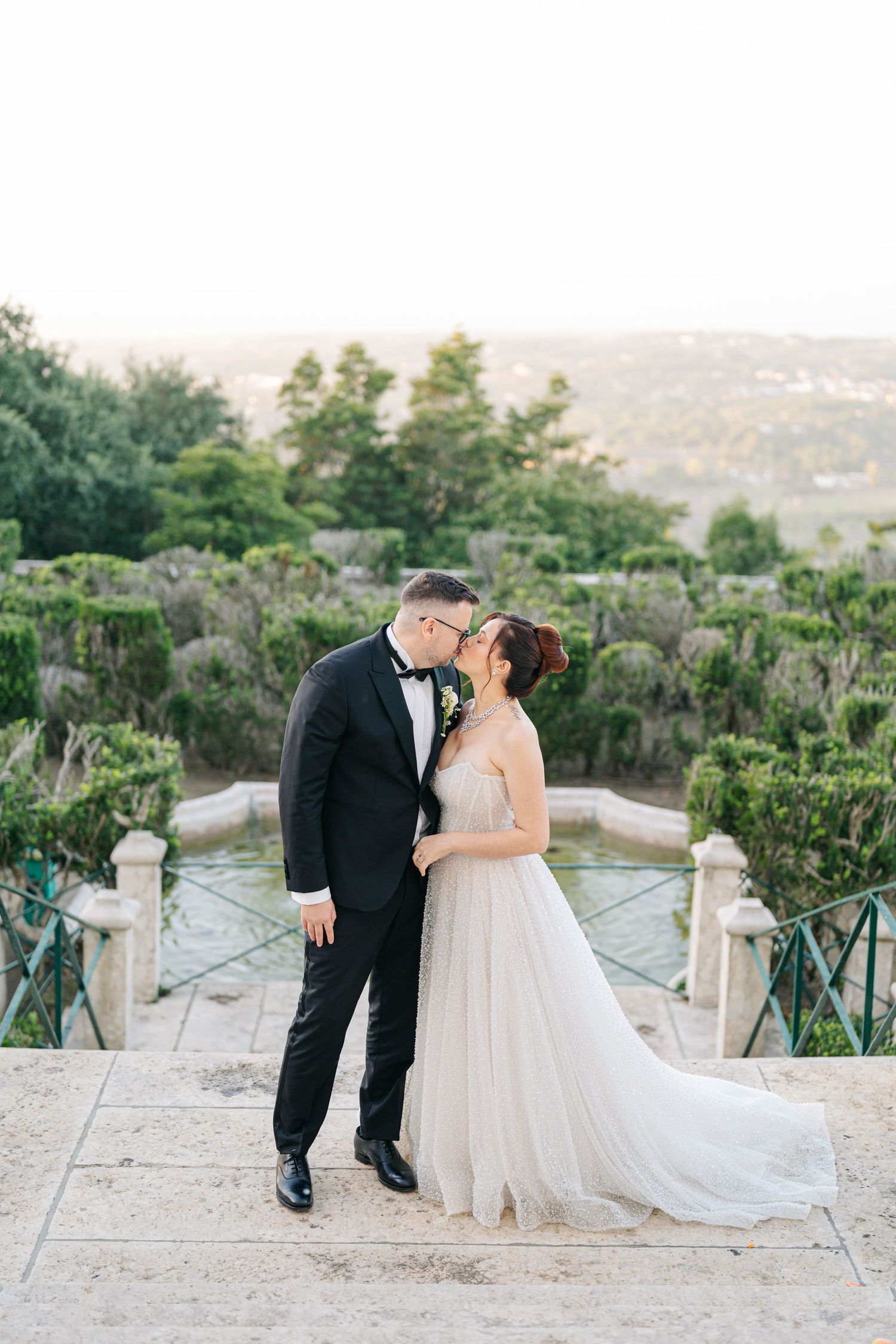 Romantic wedding couple sharing a kiss on a stone terrace overlooking scenic hillside views at sunset.