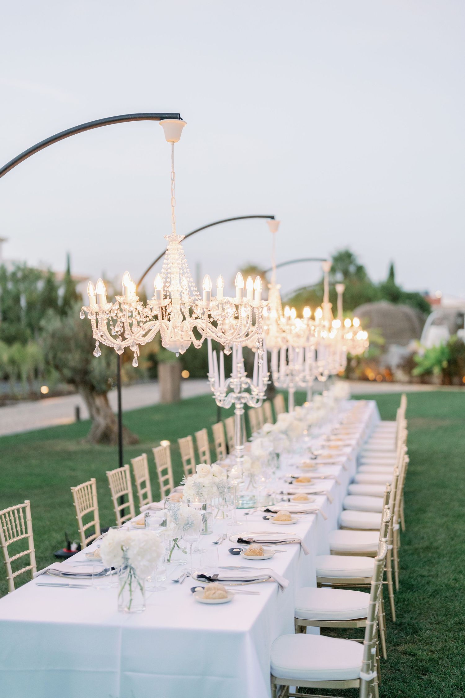 Long banquet tables with crystal chandeliers set for elegant outdoor wedding reception.