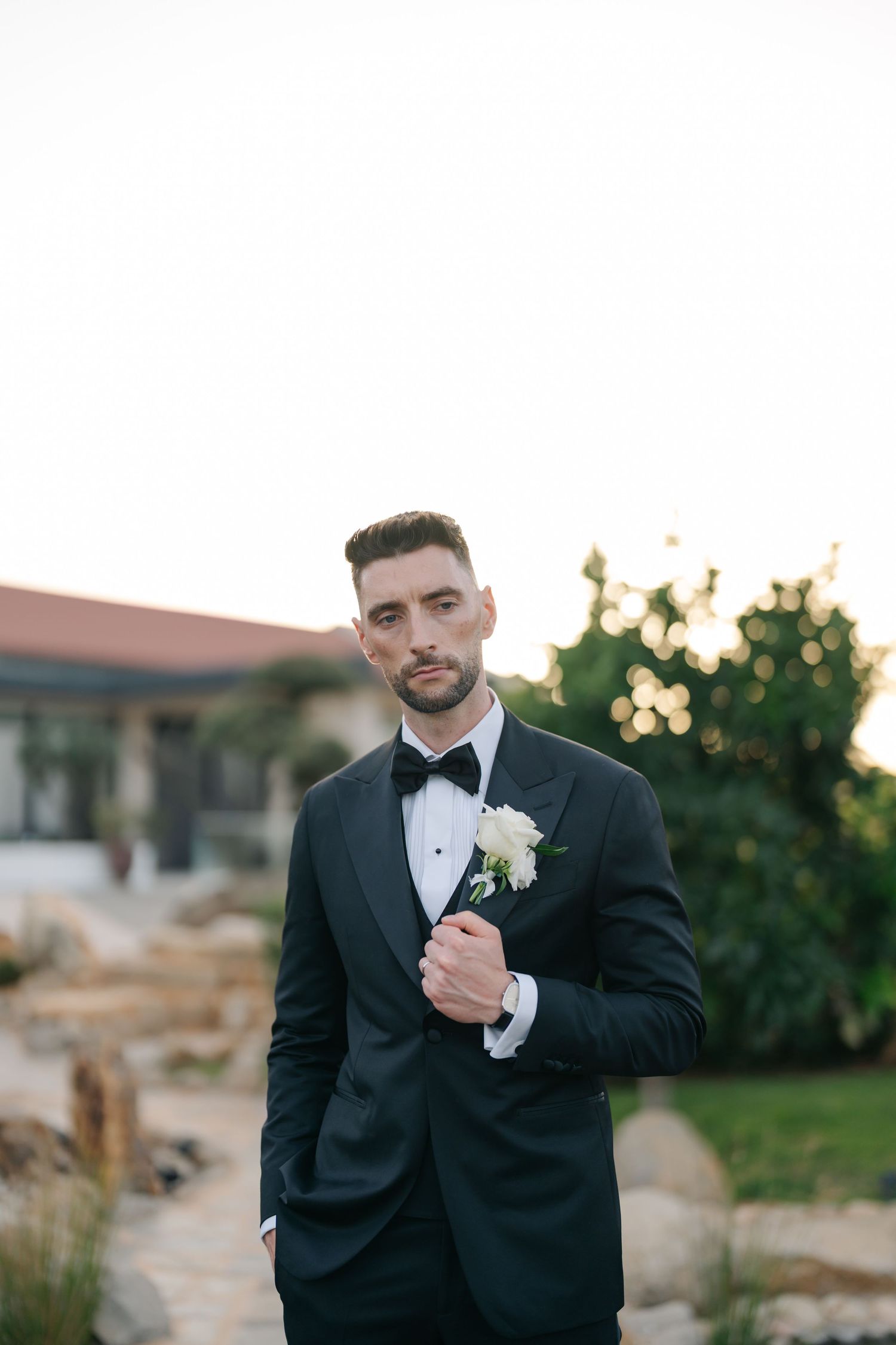 Groom in black tuxedo and bowtie poses outdoors during sunset wedding photoshoot.