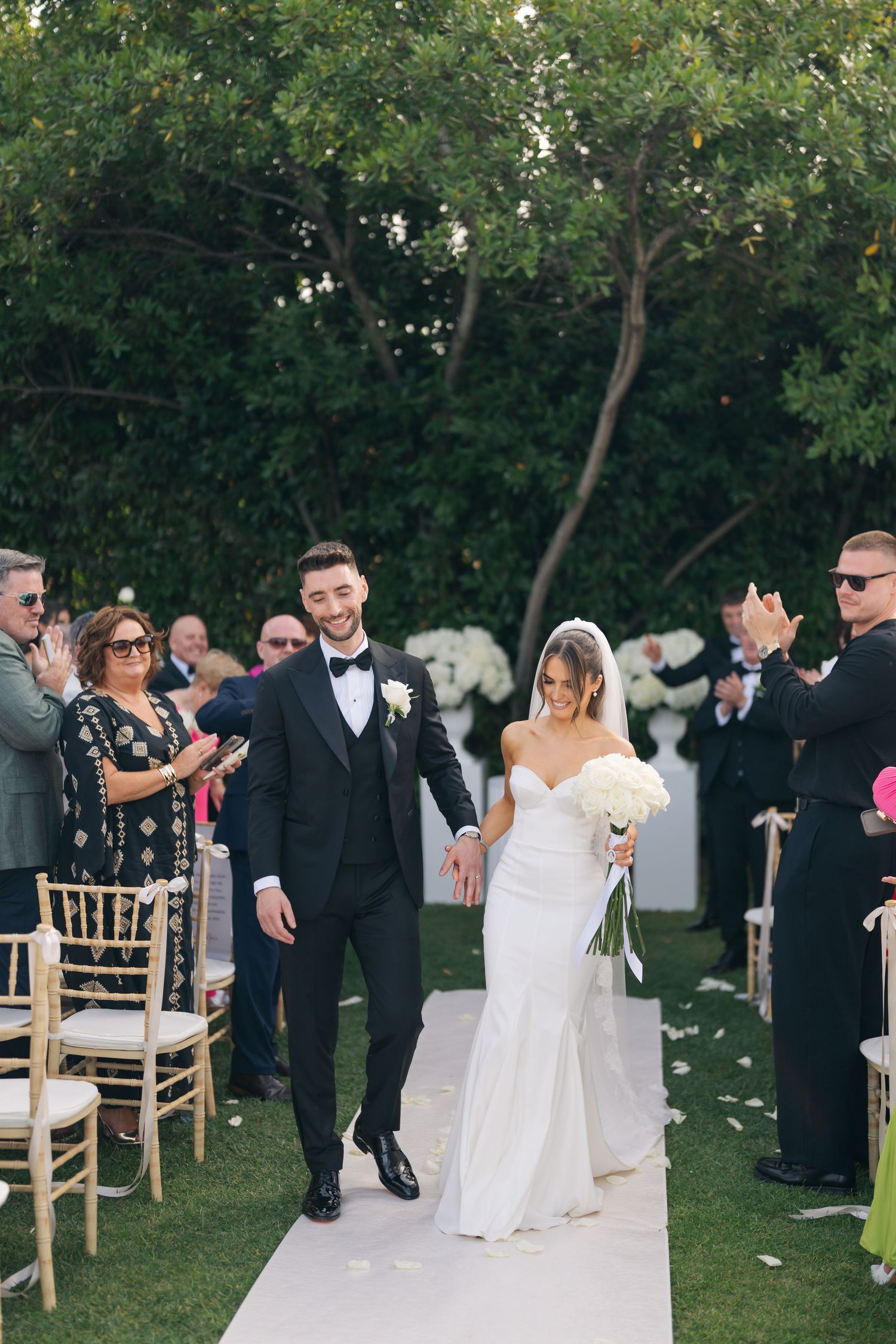 Wedding ceremony in progress on a lawn with white chairs and floral decorations.