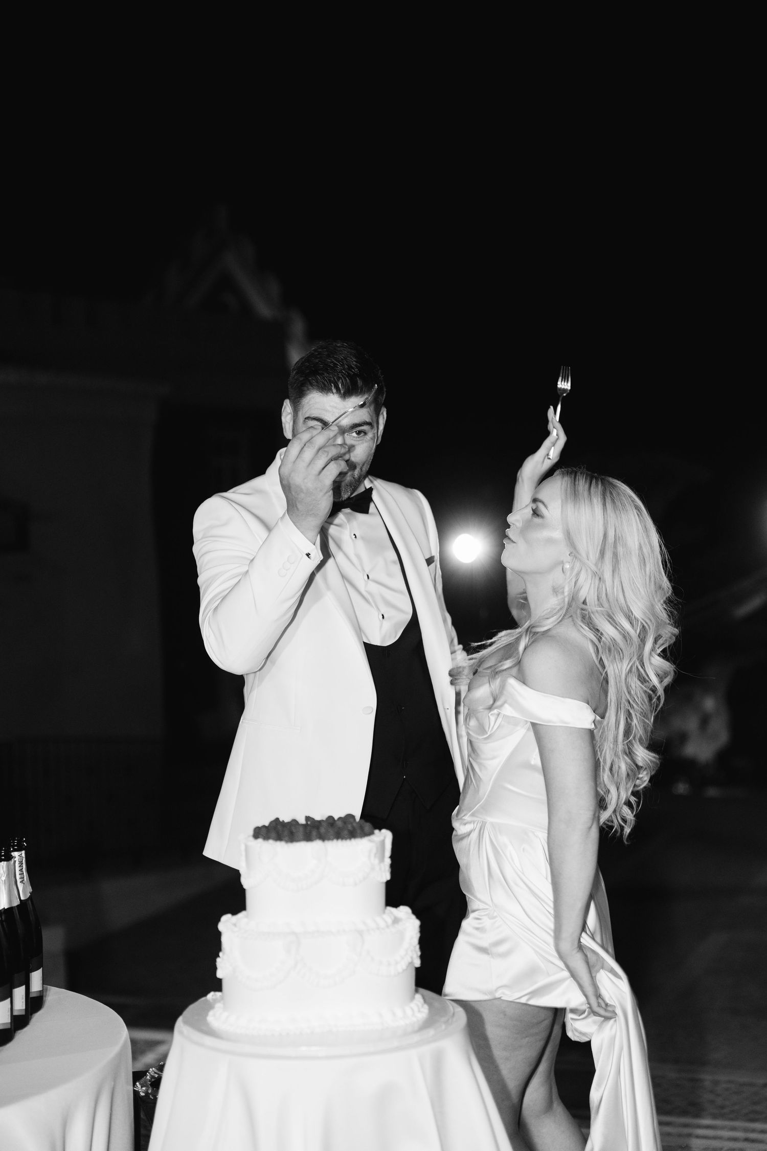 A couple in white attire celebrating near a wedding cake at night.