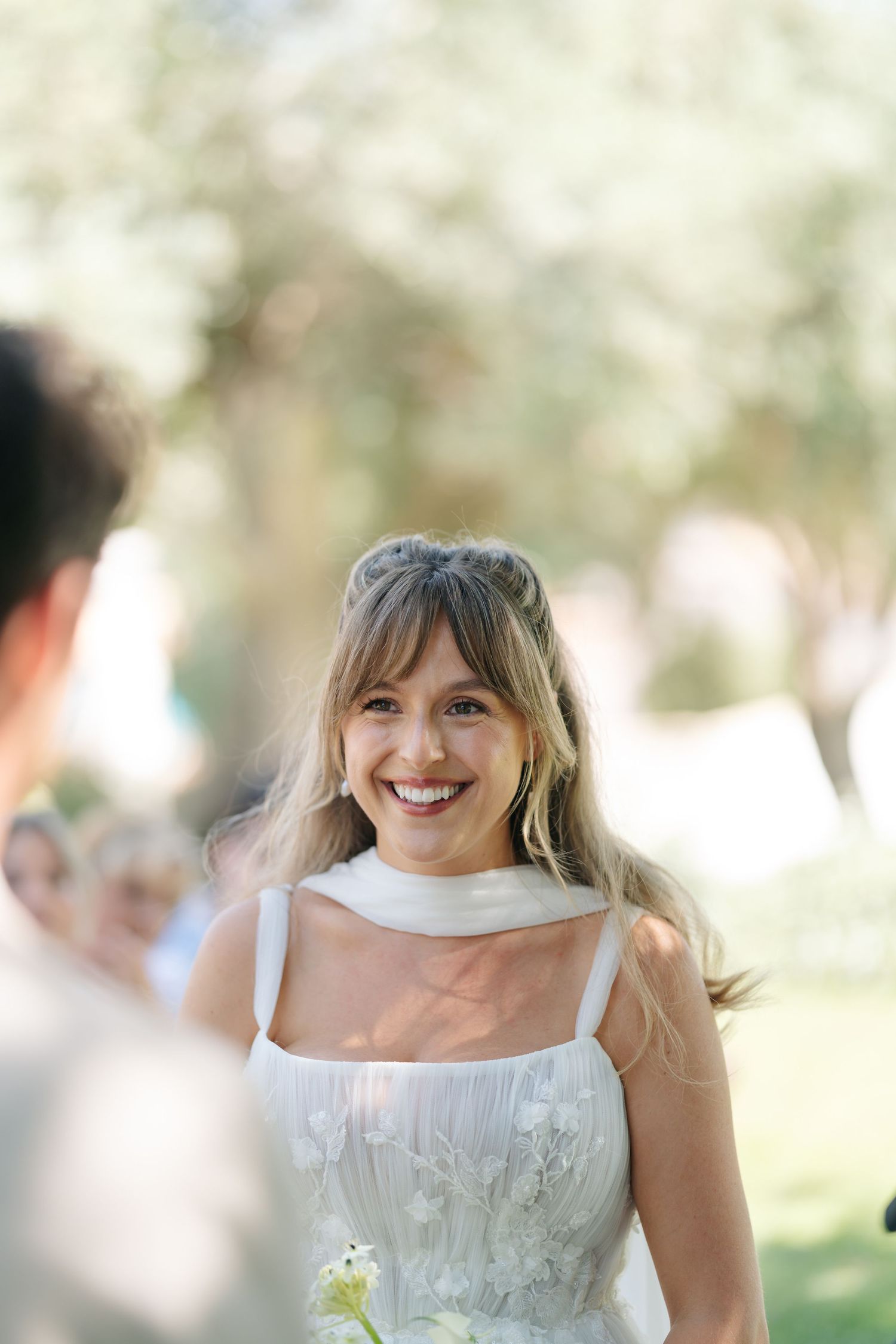 A bride in a white wedding dress smiles joyfully in an outdoor garden setting with soft bokeh lighting effects.