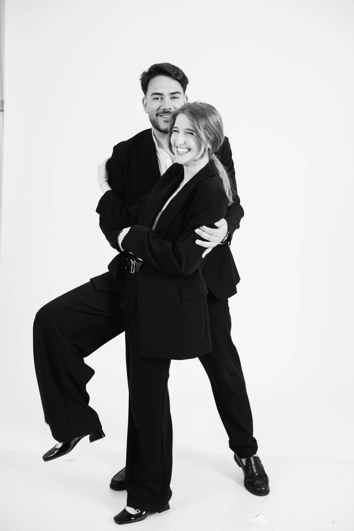 Three black and white photos show two people in dark suits playfully posing together against a white backdrop.