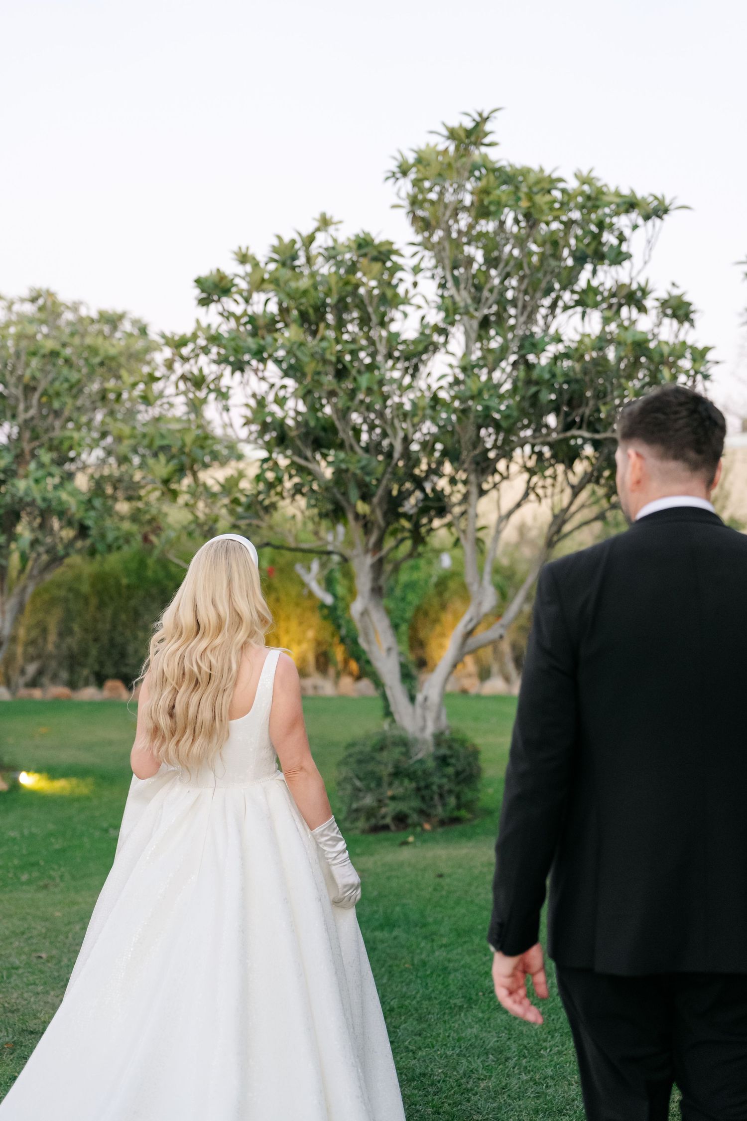 A couple in wedding attire strolls through a scenic garden with lush greenery.