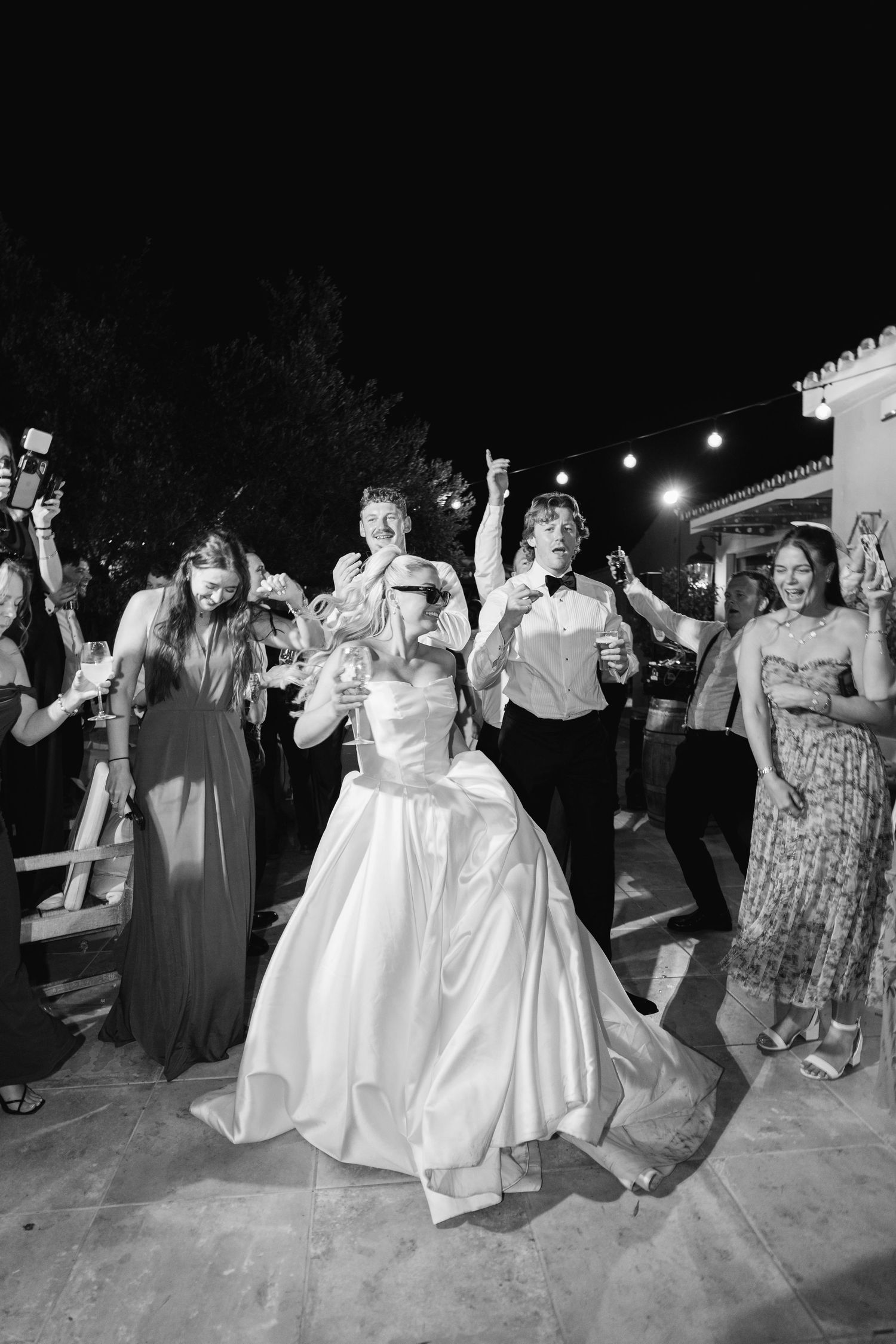 Wedding guests dancing in a line at an outdoor reception with string lights overhead.