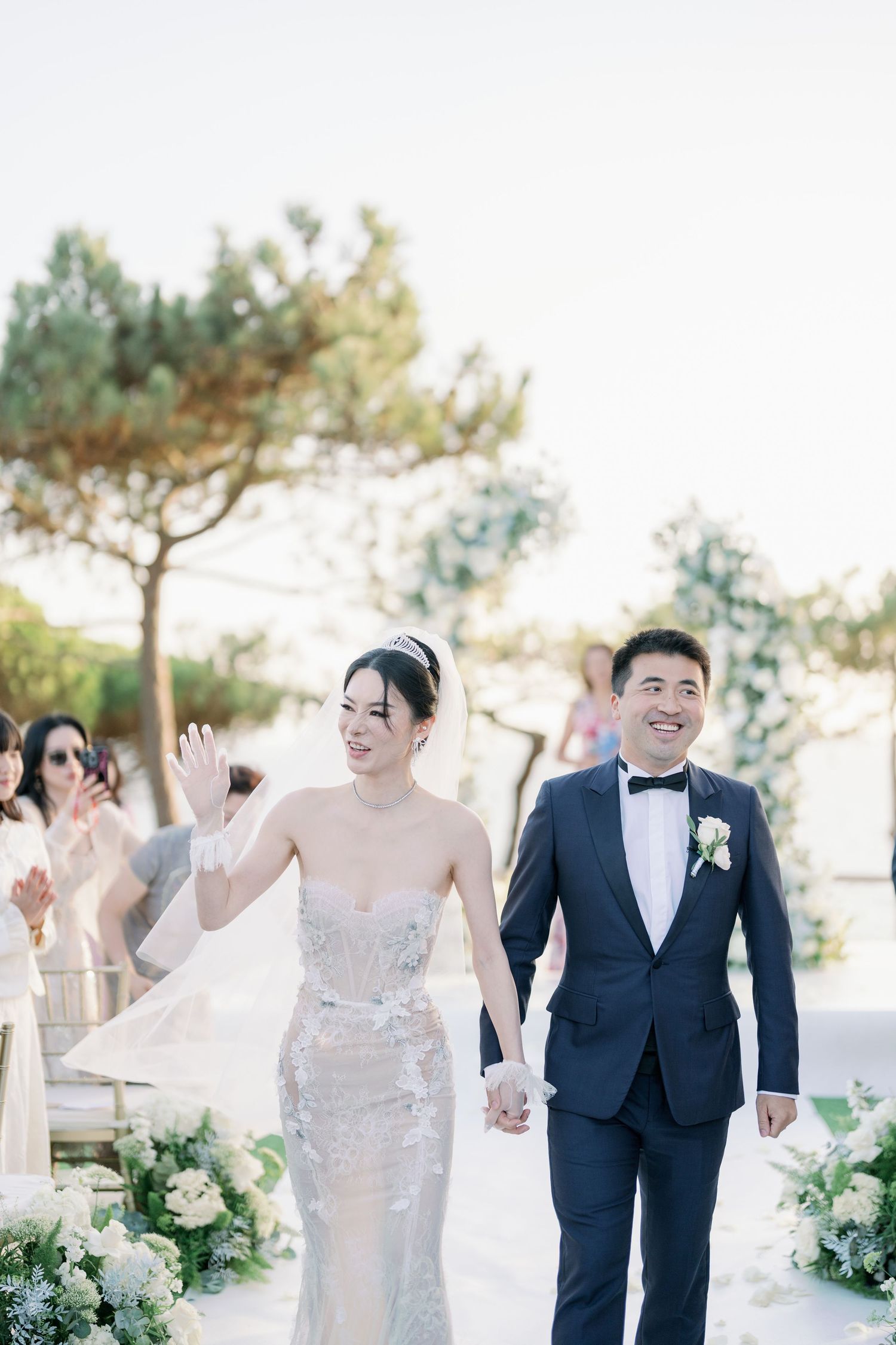 Newlyweds walk down the aisle together surrounded by white floral decorations at an outdoor wedding ceremony.
