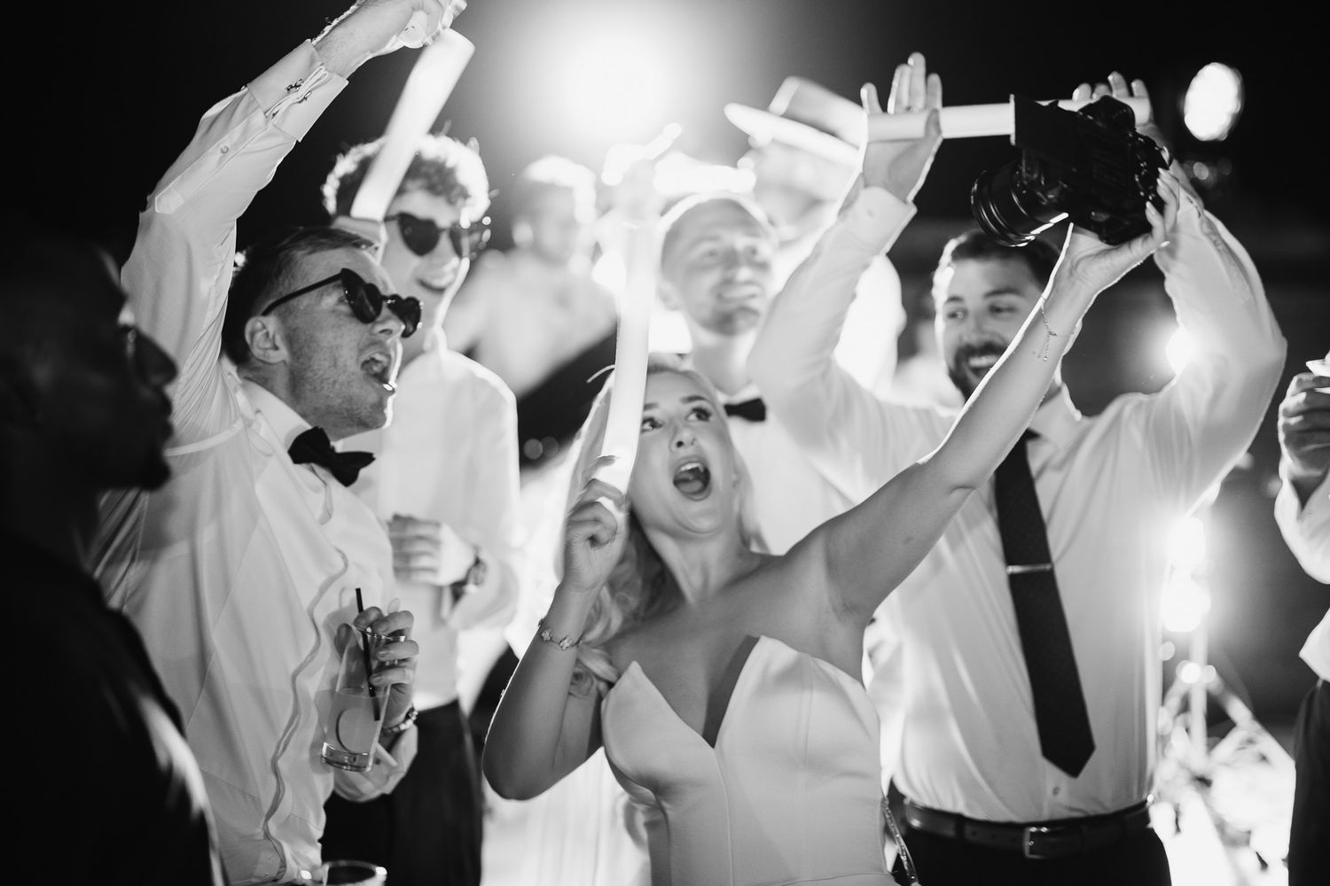 Wedding guests dancing and celebrating with raised arms in bright lighting.