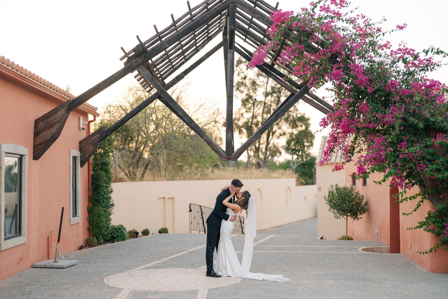 A romantic wedding couple poses under pink bougainvillea flowers at an outdoor venue.