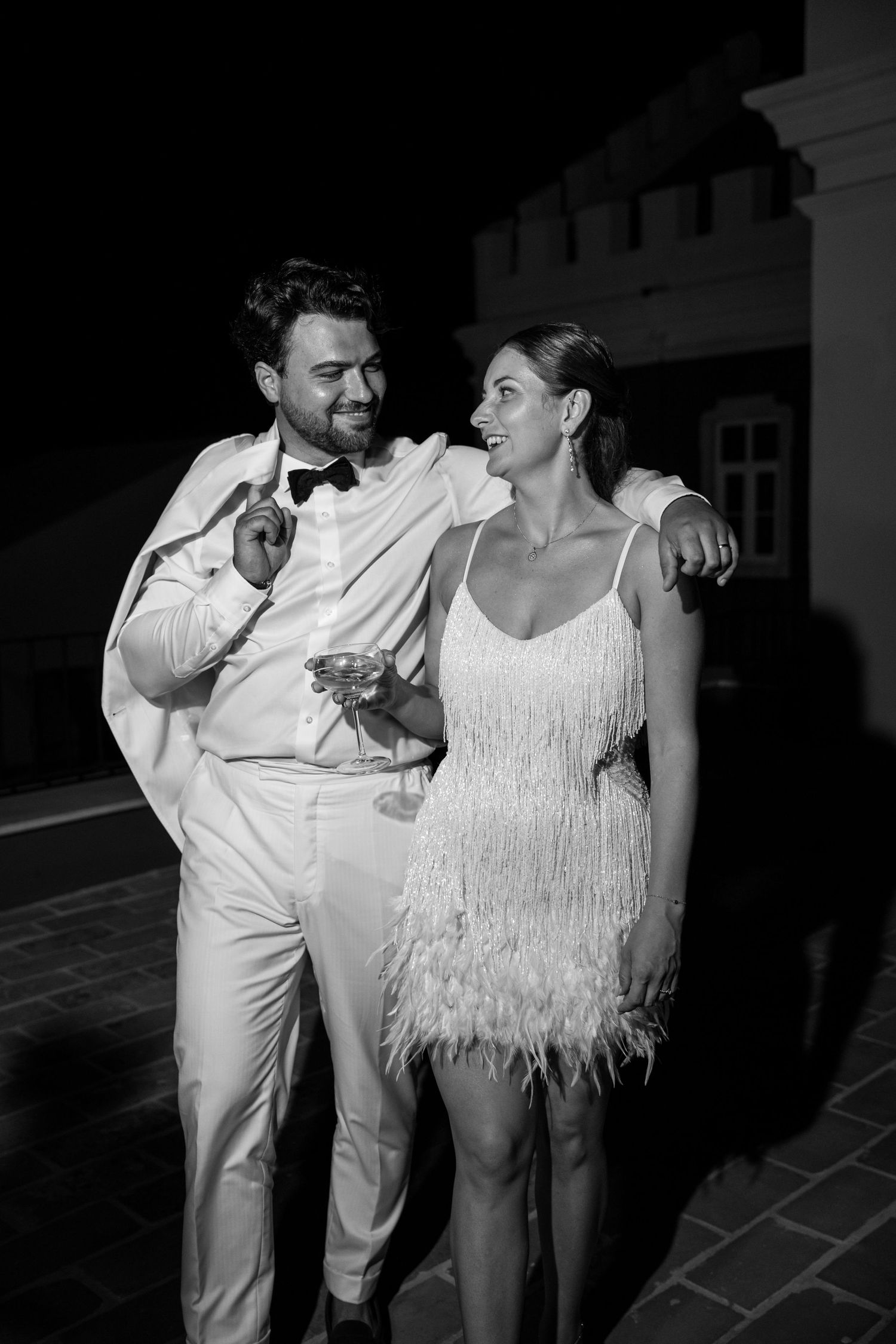 A couple shares a joyful moment at a wedding reception, with the bride wearing a white fringe dress and groom in white suit.