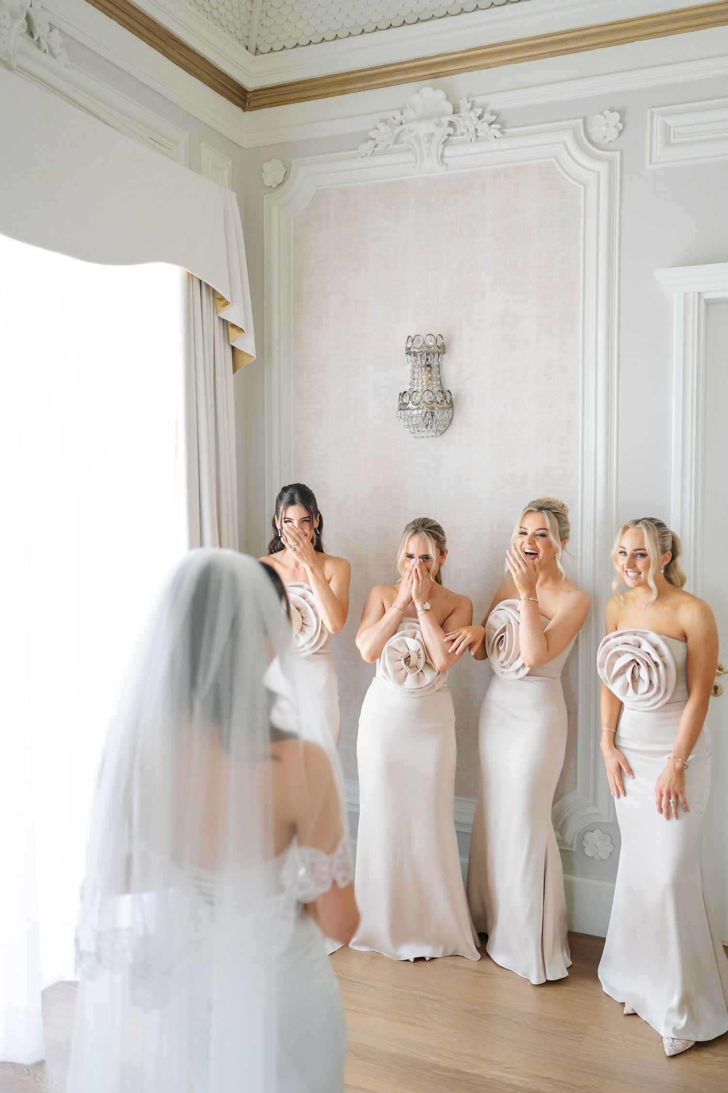 Bridesmaids in white dresses react to seeing bride in wedding attire.