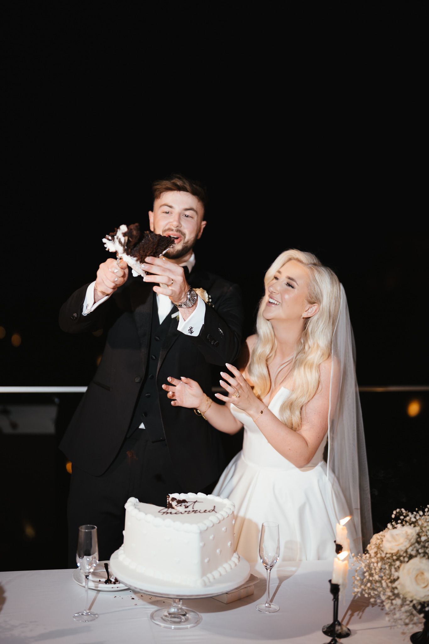Couple shares joyful moment while cutting their white wedding cake at evening reception.