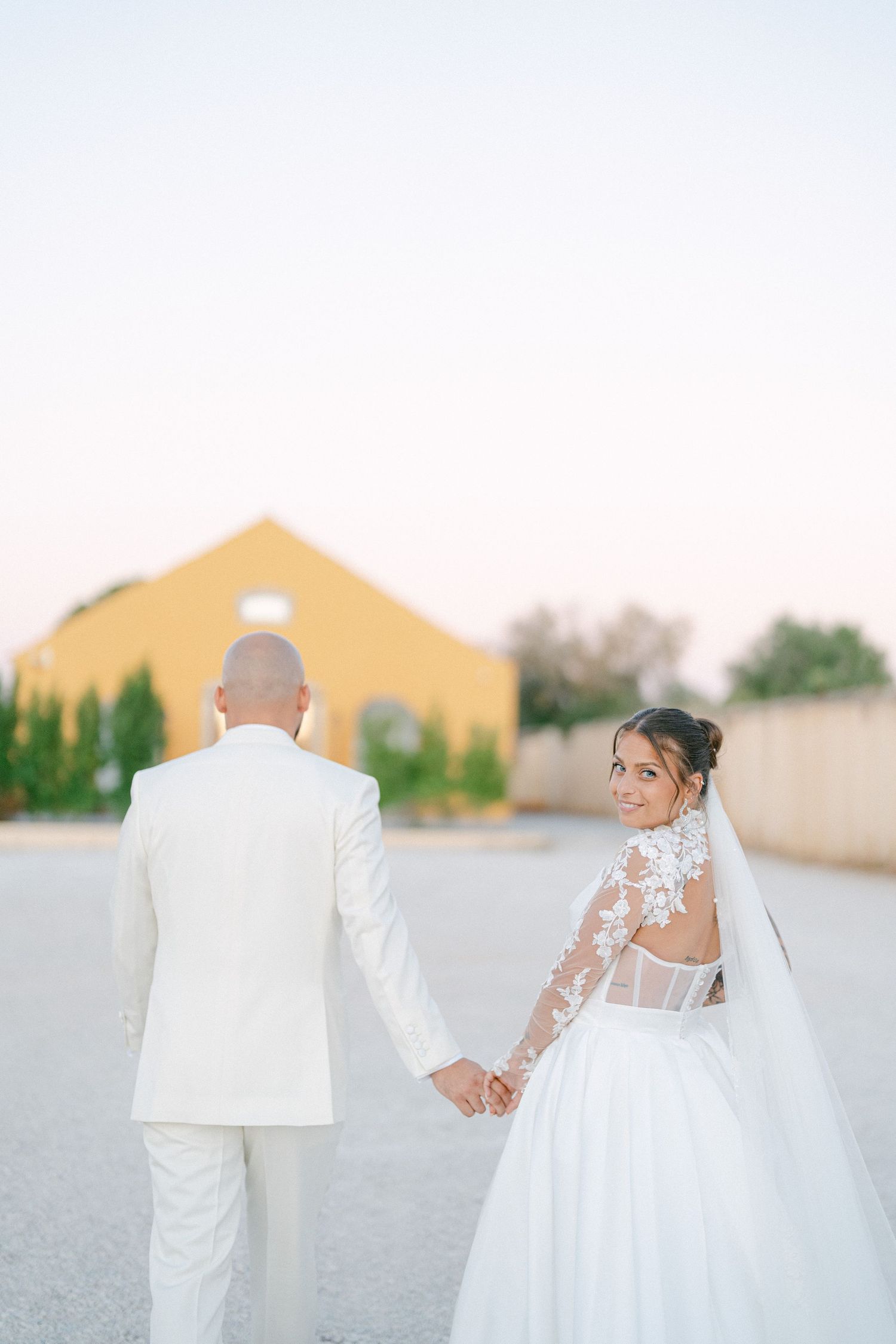 A couple in white wedding attire walks together on a path with a yellow building in the background.