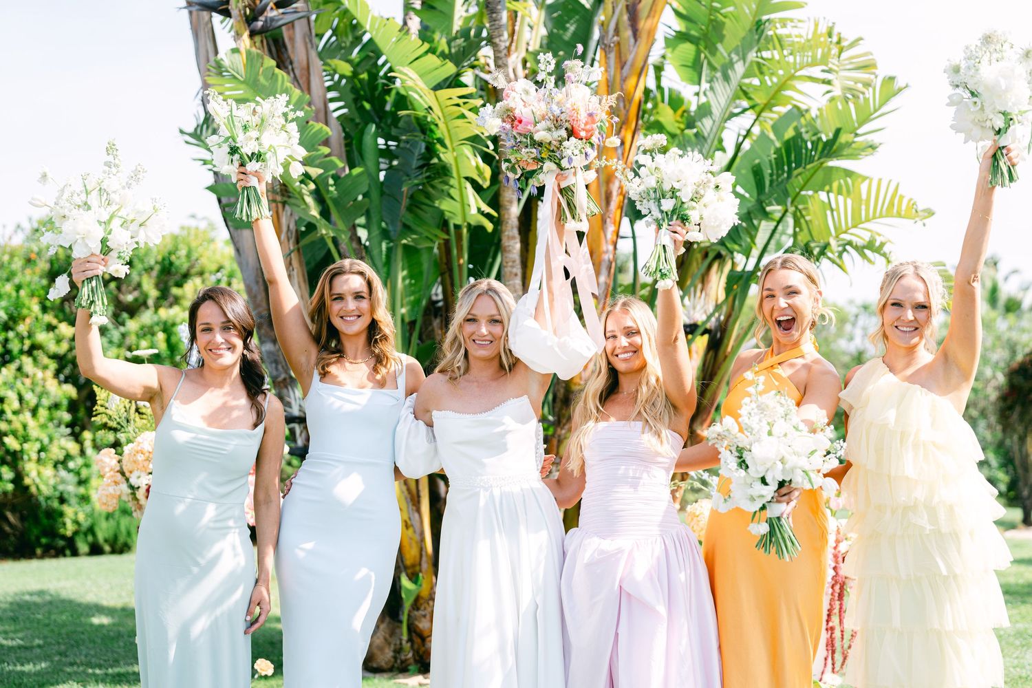 A group in white and pastel dresses poses together in a lush garden setting for a summer celebration.