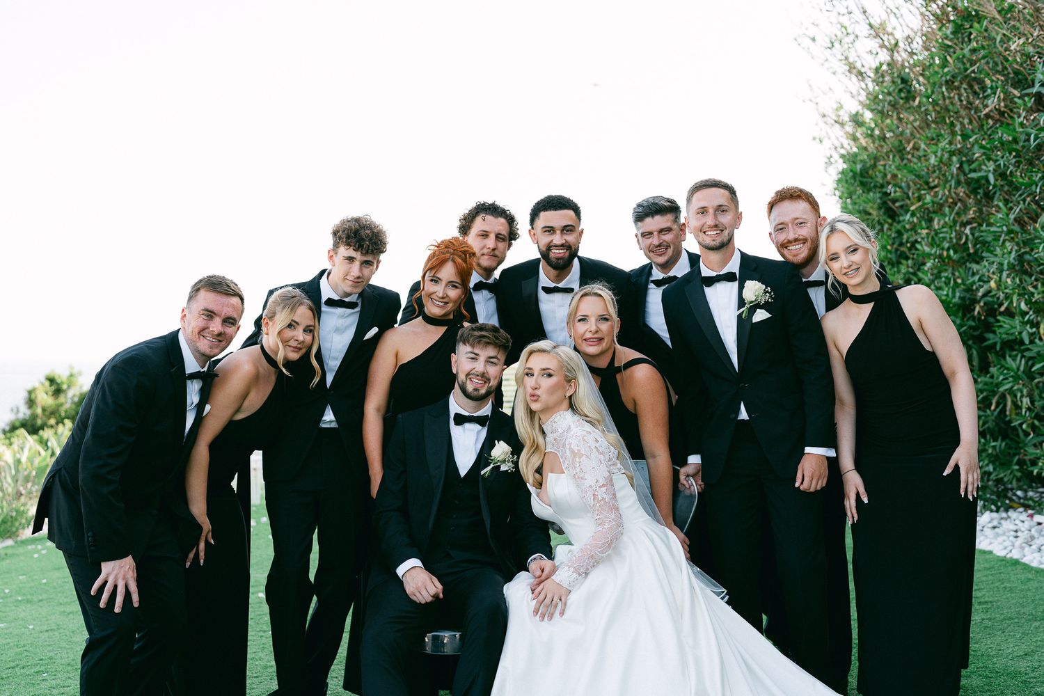 Wedding party poses playfully on grassy lawn in formal attire with black suits and white gowns.