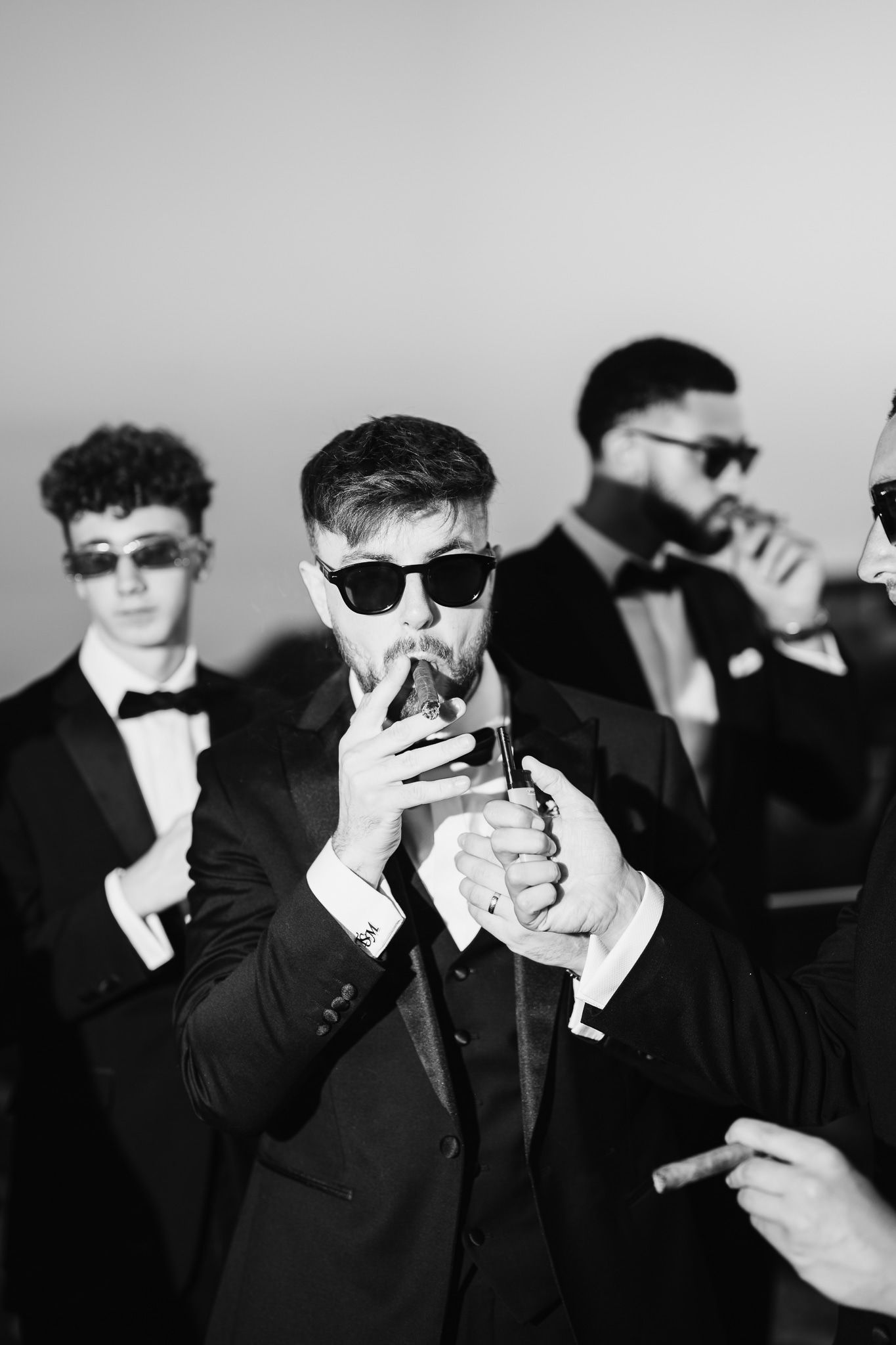 Wedding party members in formal attire and sunglasses light cigarettes in dramatic black and white.
